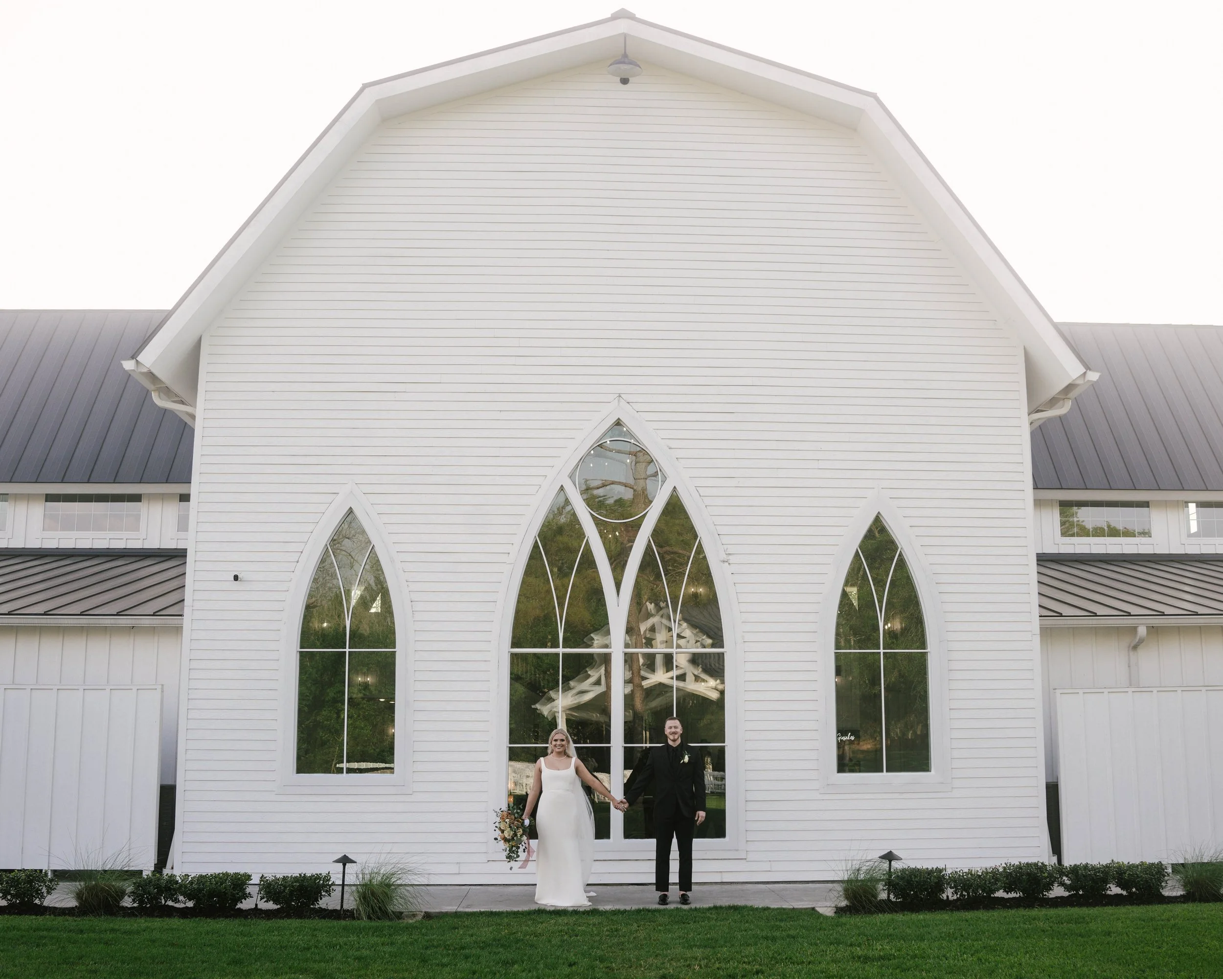 A bride and groom holding hands and smiling in front of a white church with large pointed stained glass windows, greenery, and a manicured lawn.