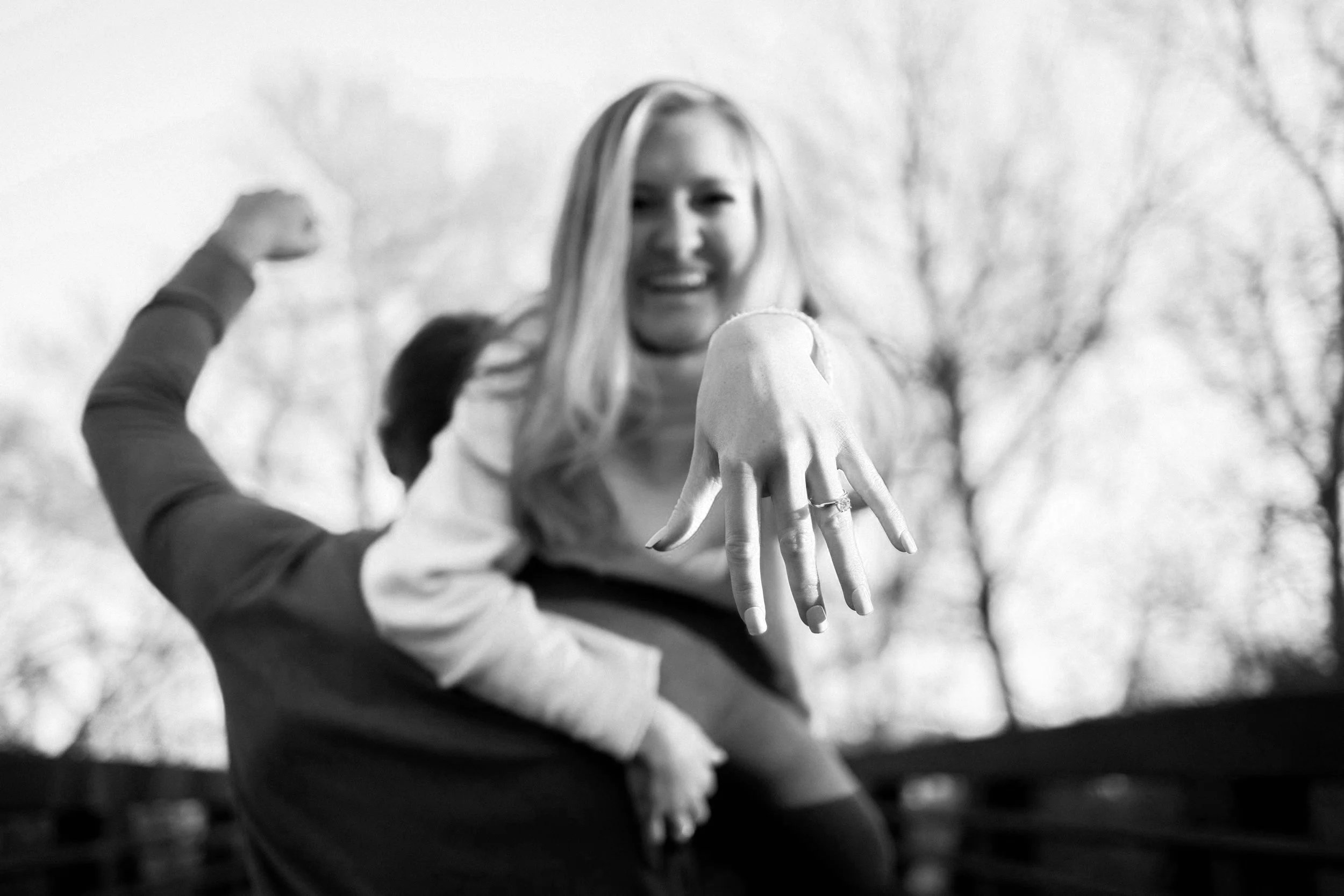 A woman with long blonde hair showing her hand with rings, with a smiling man flexing his arm in the background, outdoors with leafless trees.