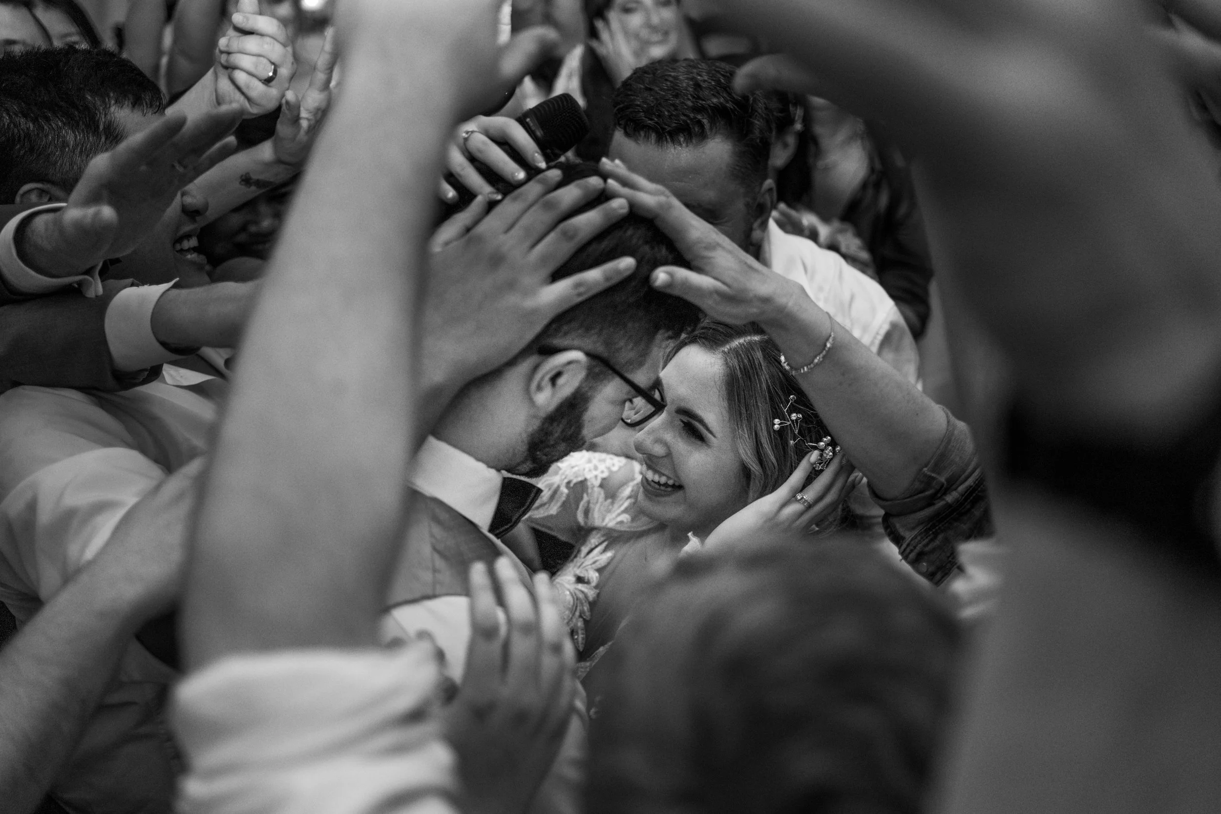 A black and white photo of a wedding celebration showing a bride and groom close together, surrounded by friends and family who are reaching out and touching their heads and shoulders, capturing a joyful and intimate moment.