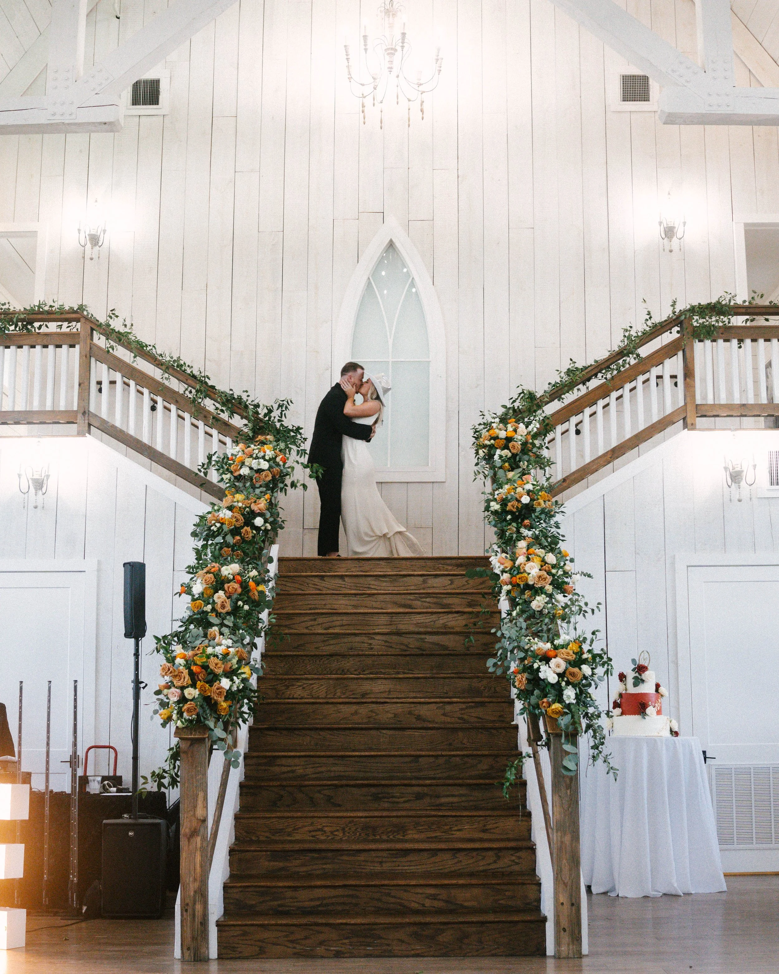 A bride and groom sharing a kiss on a staircase decorated with floral arrangements in a white church with wooden accents.