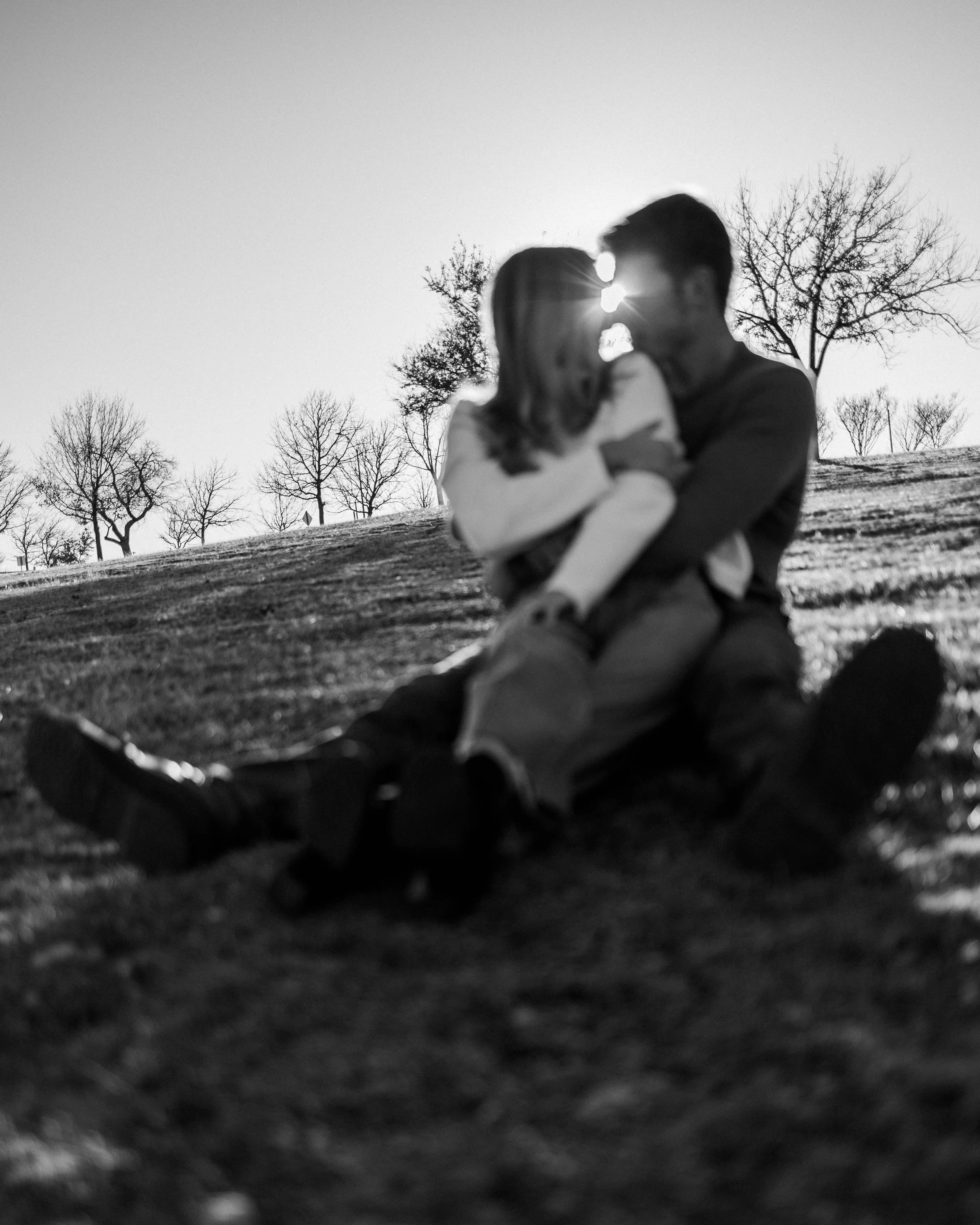 A black and white photo of a couple sitting on the grass, kissing, with trees and the sun in the background.