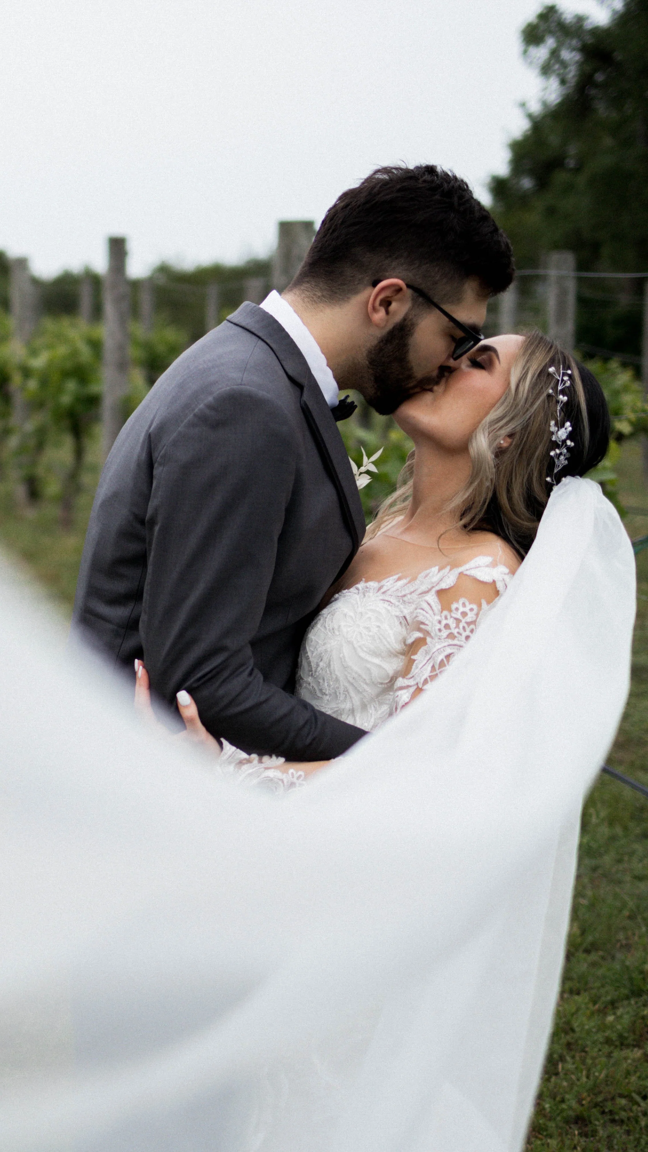 A bride and groom kiss outdoors, with the groom wearing a gray suit and the bride in a white lace wedding dress, standing in a green vineyard.