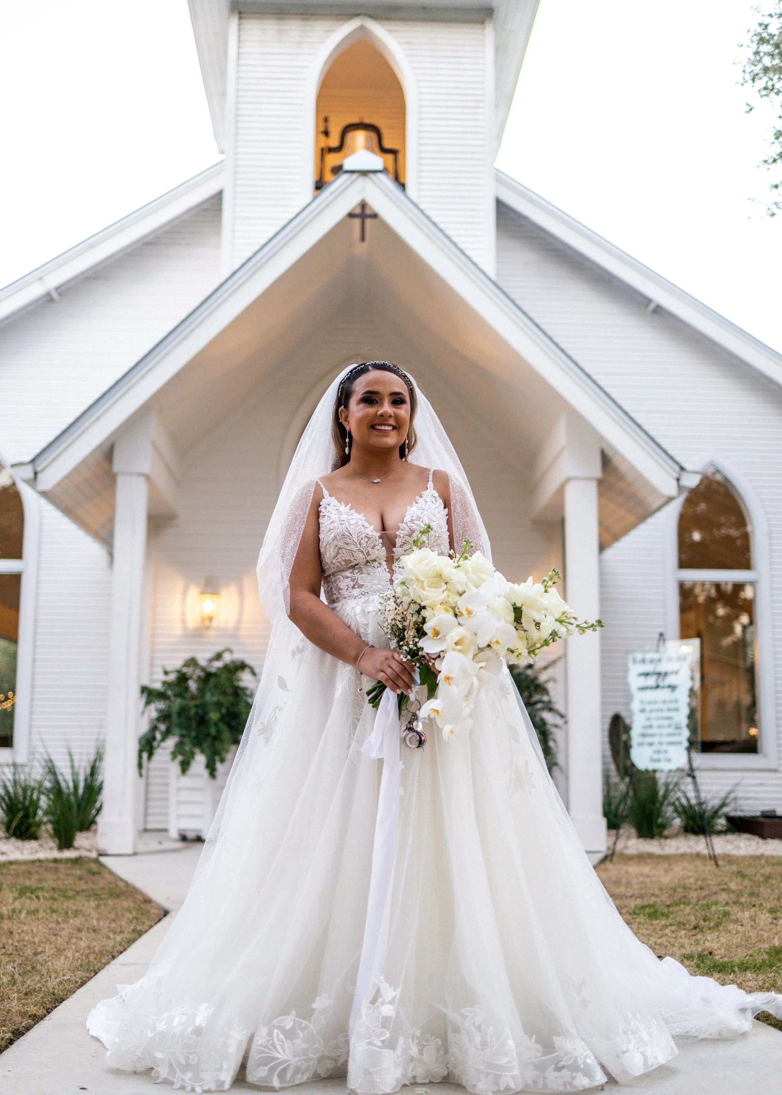 A bride in a white wedding gown standing outside a white church with a steeple, holding a bouquet of white flowers, and smiling.