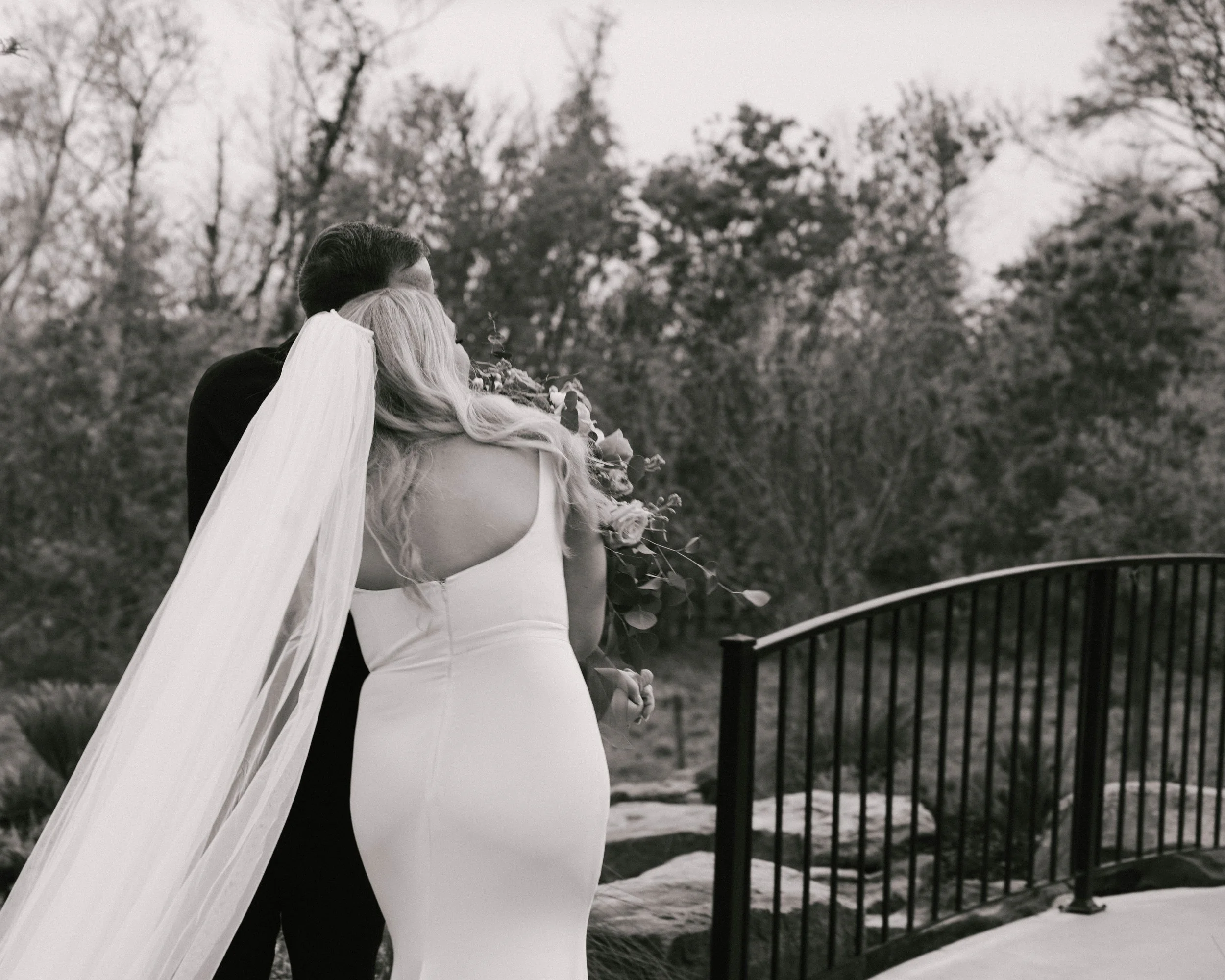 A bride and groom embrace outdoors on a stone path with trees in the background, black and white photo.