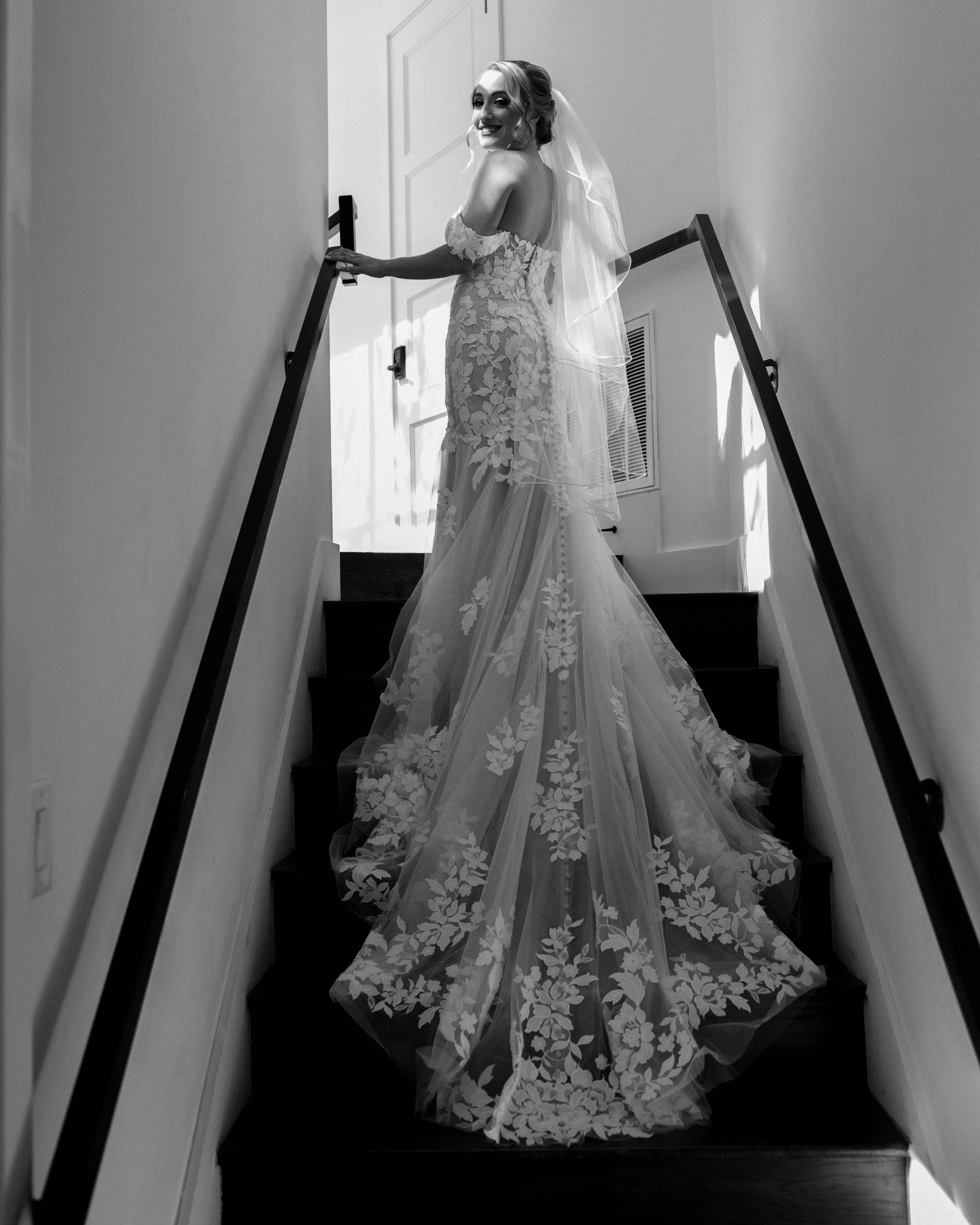 A bride in a lace wedding dress with off-the-shoulder sleeves and a long train, smiling while standing on carpeted stairs indoors.