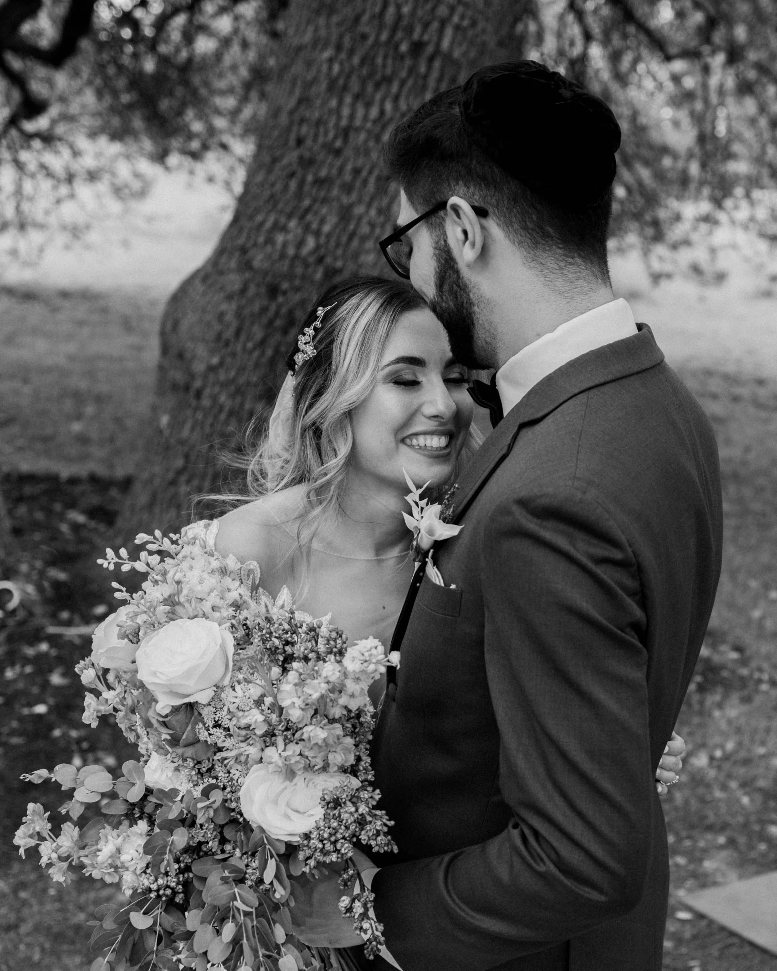 A black and white photo of a smiling bride holding a bouquet and a groom facing her with their foreheads touching, outdoors near a tree.