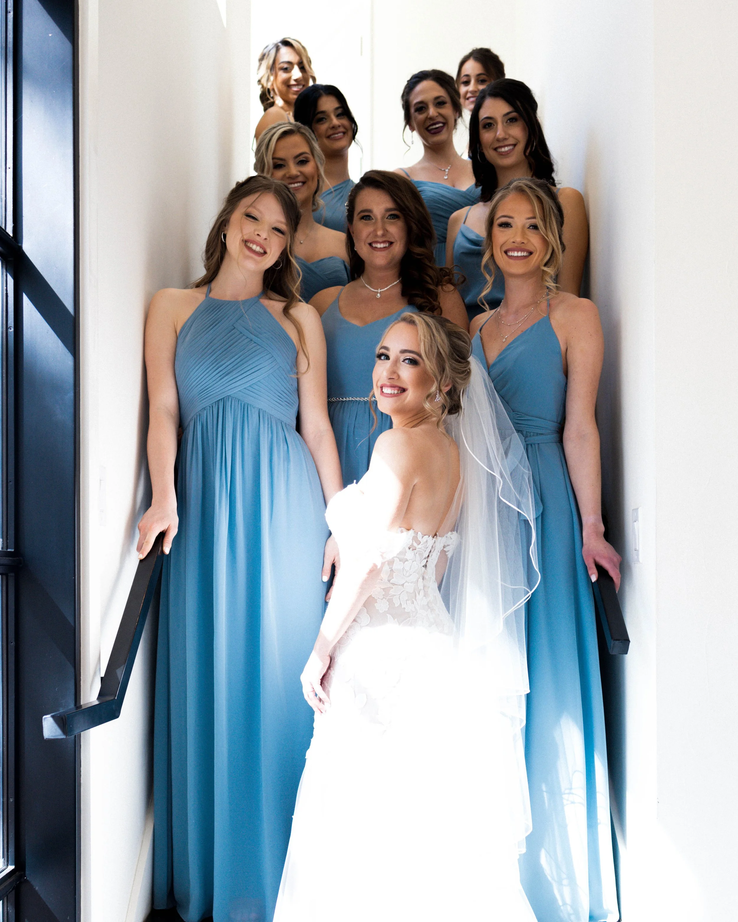 Bride in a white wedding dress with veil and ten bridesmaids in matching blue dresses standing on a staircase.