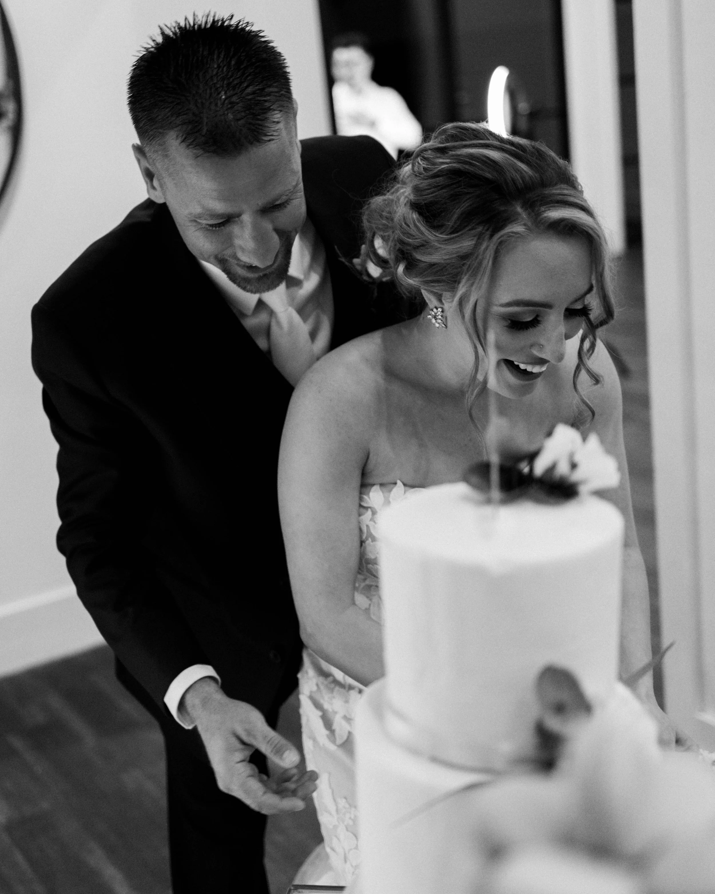 A black and white photo of a bride and groom cutting their wedding cake together, smiling and leaning close.