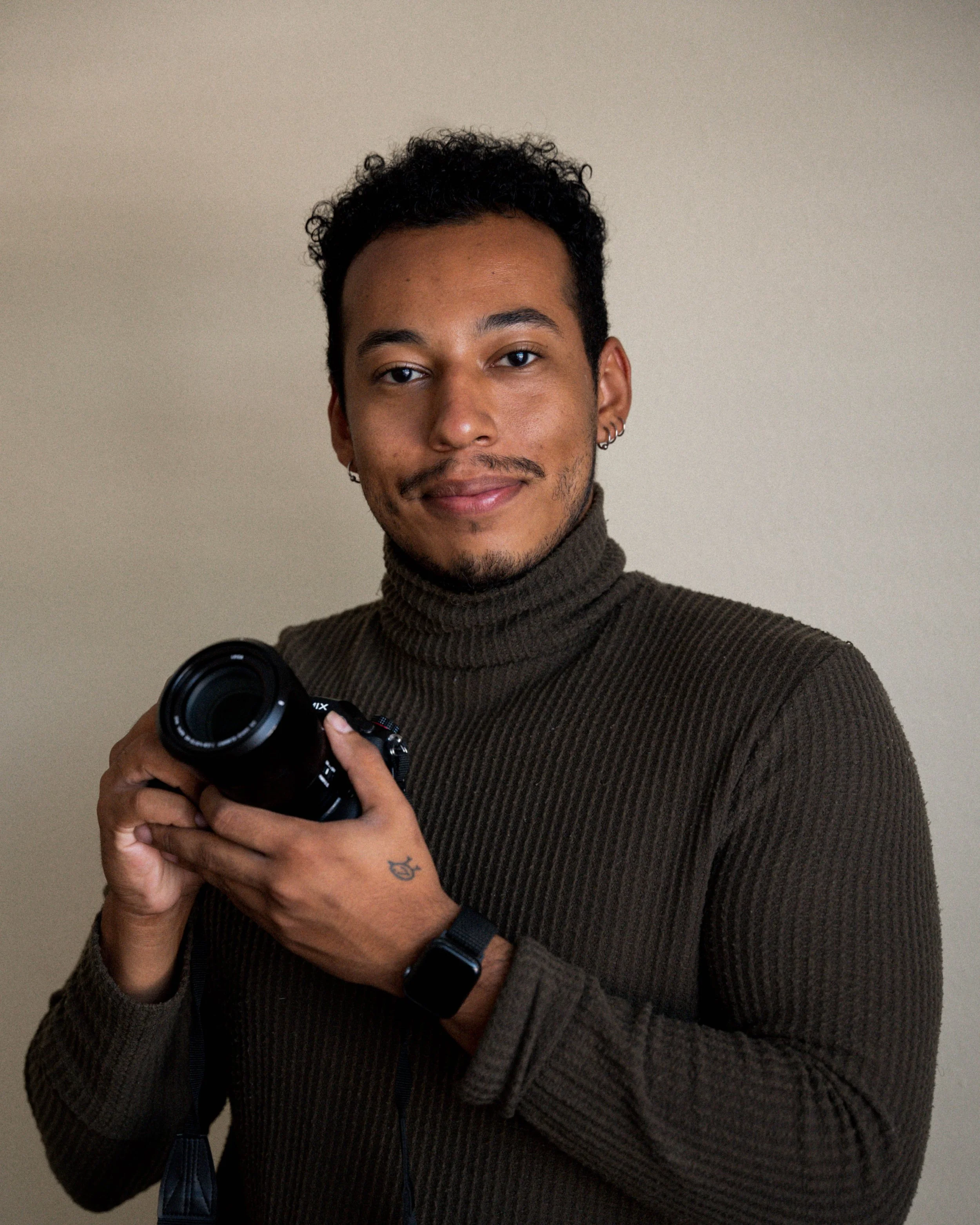 A young man with curly hair, earrings, and a goatee, wearing a brown ribbed turtleneck sweater, holding a camera and smiling. He is standing against a plain beige background.