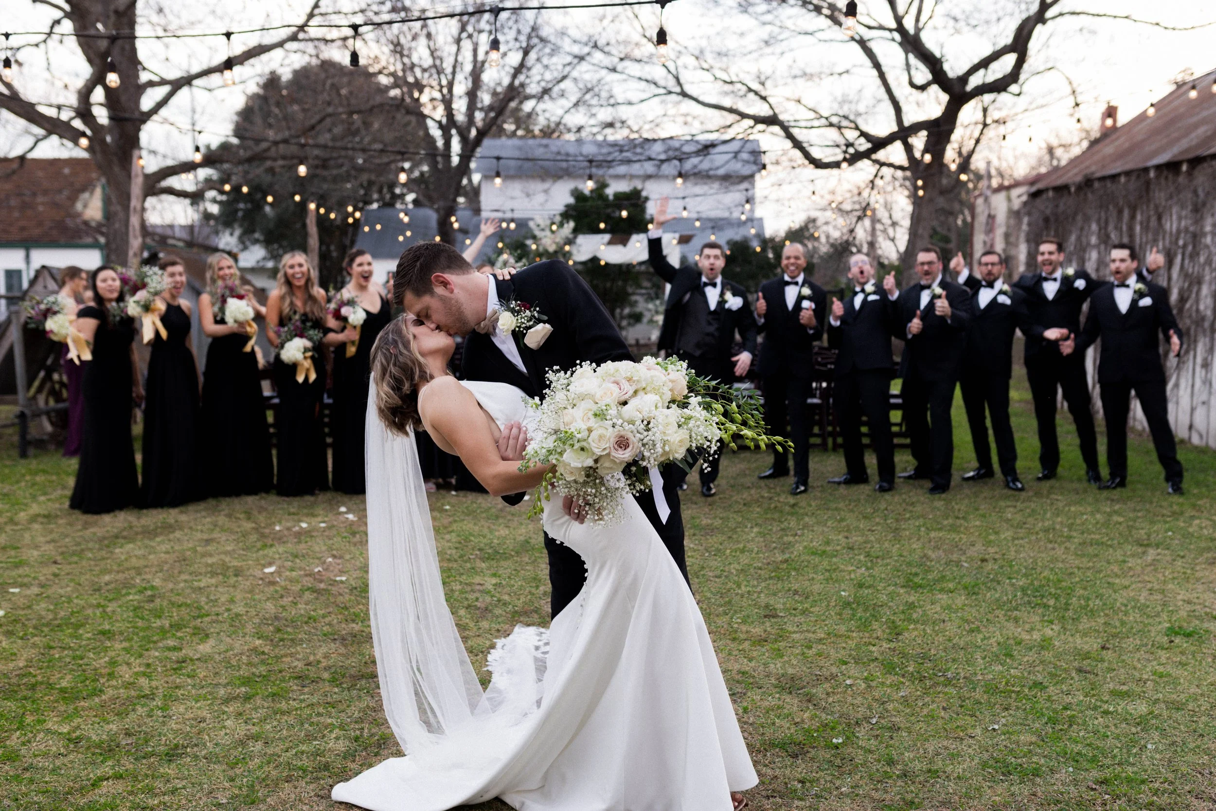A newlywed couple sharing a kiss, with the groom dipping the bride, in front of their wedding party outdoors at sunset. The bridesmaids and groomsmen are standing in a line behind them, dressed in black and tuxedos, surrounded by trees with string lights hanging overhead.