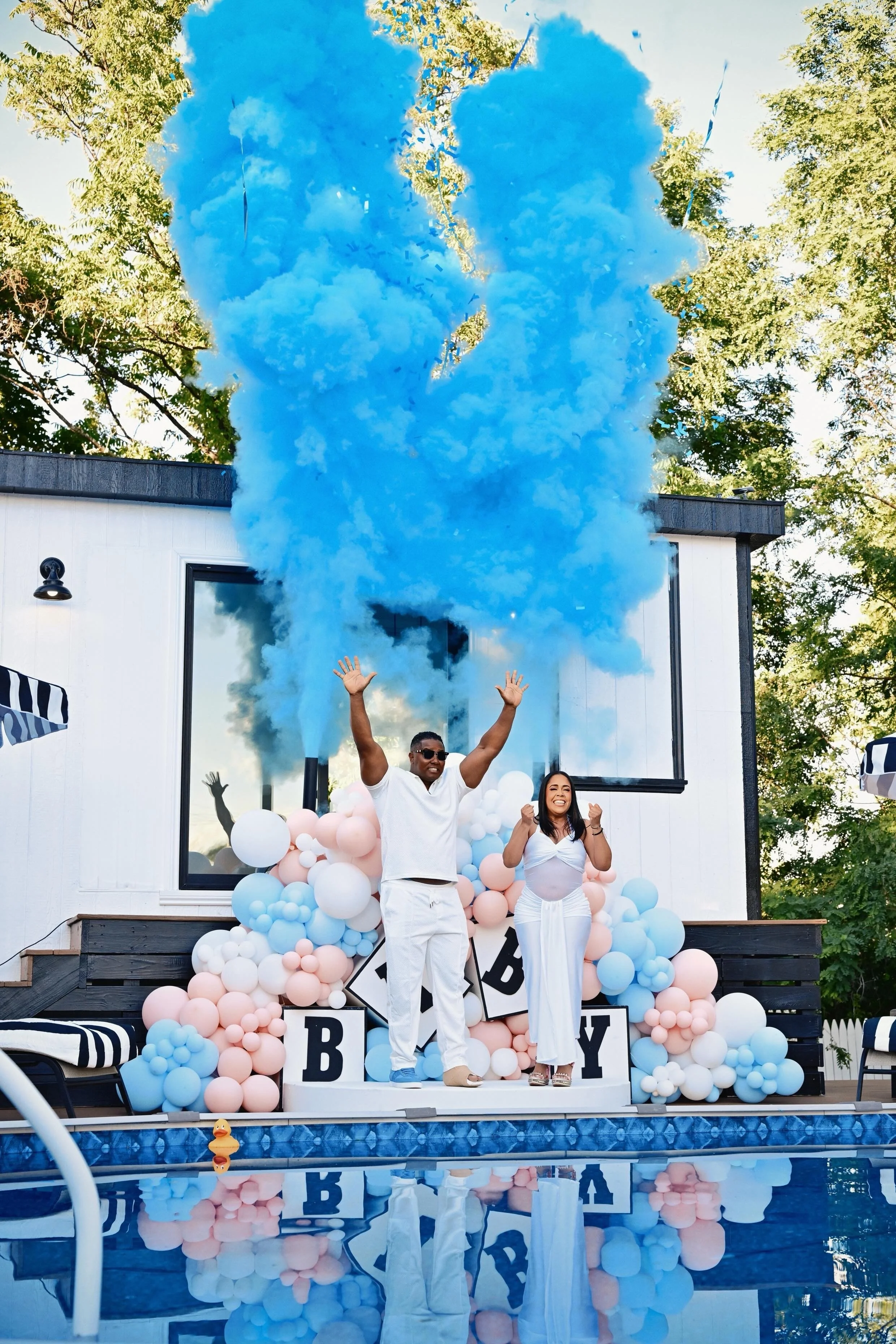 Expectant couple celebrating a baby boy gender reveal beside a pool with blue smoke, confetti, and pink, blue, and white balloons.