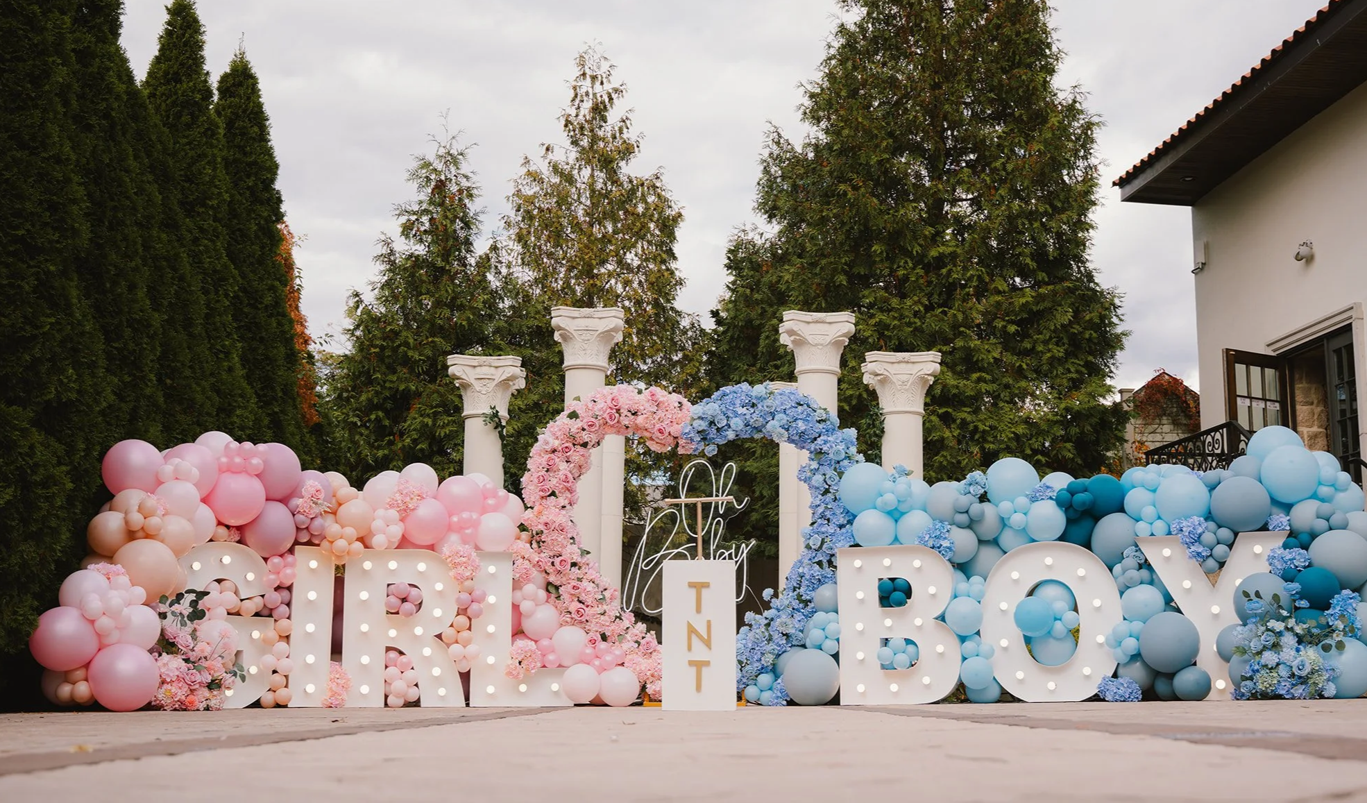 Outdoor gender reveal display with illuminated GIRL and BOY marquee letters, pink and blue balloon garlands, matching floral heart arch, half pink, half blue, and a central “Oh Baby” sign.