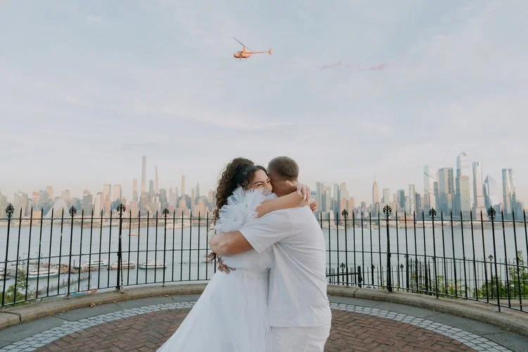 Expectant couple embracing during a waterfront helicopter gender reveal with a city skyline in the background.