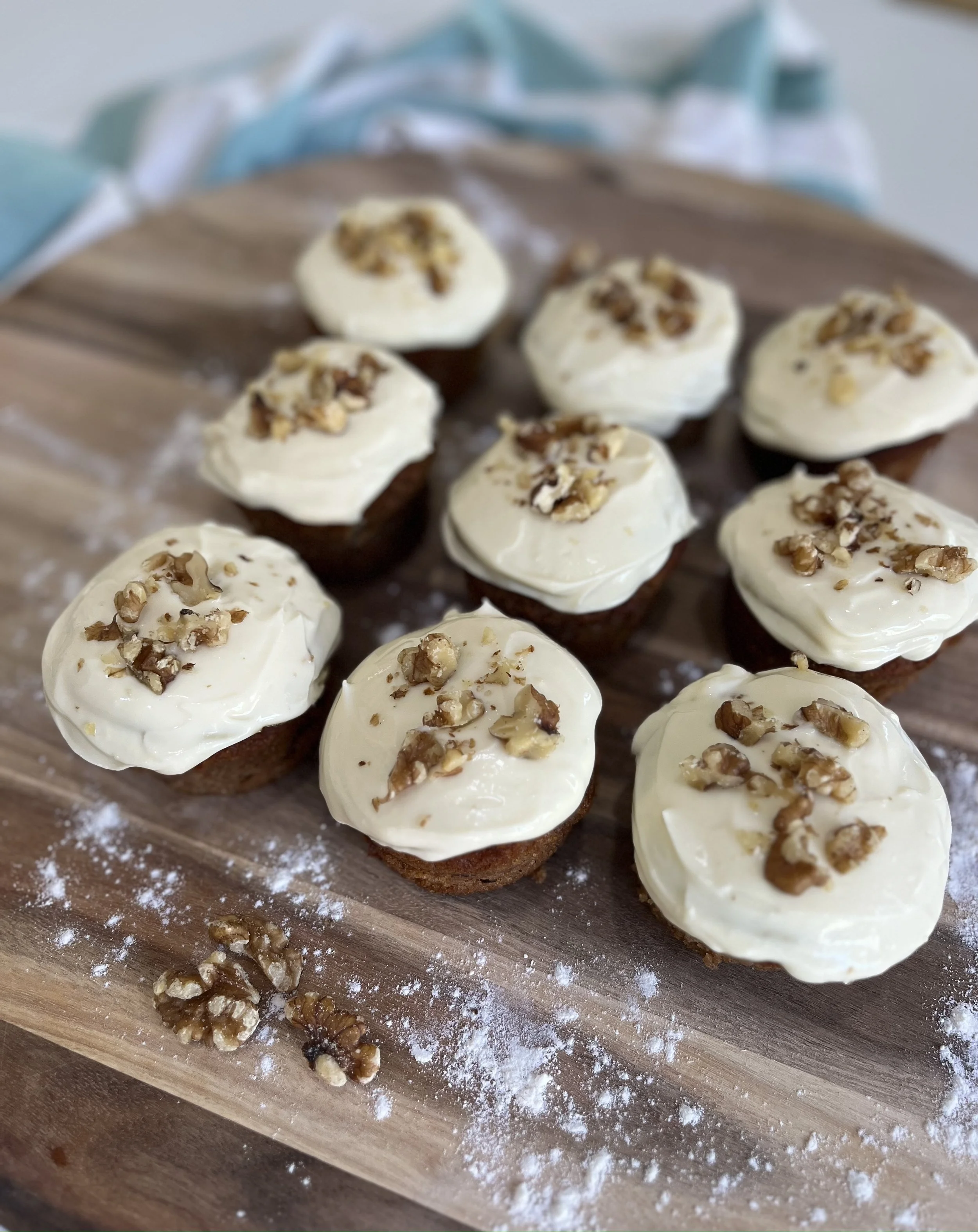 Nine cupcakes topped with white frosting and chopped walnuts, arranged on a wooden cutting board with some scattered walnuts and powdered sugar.