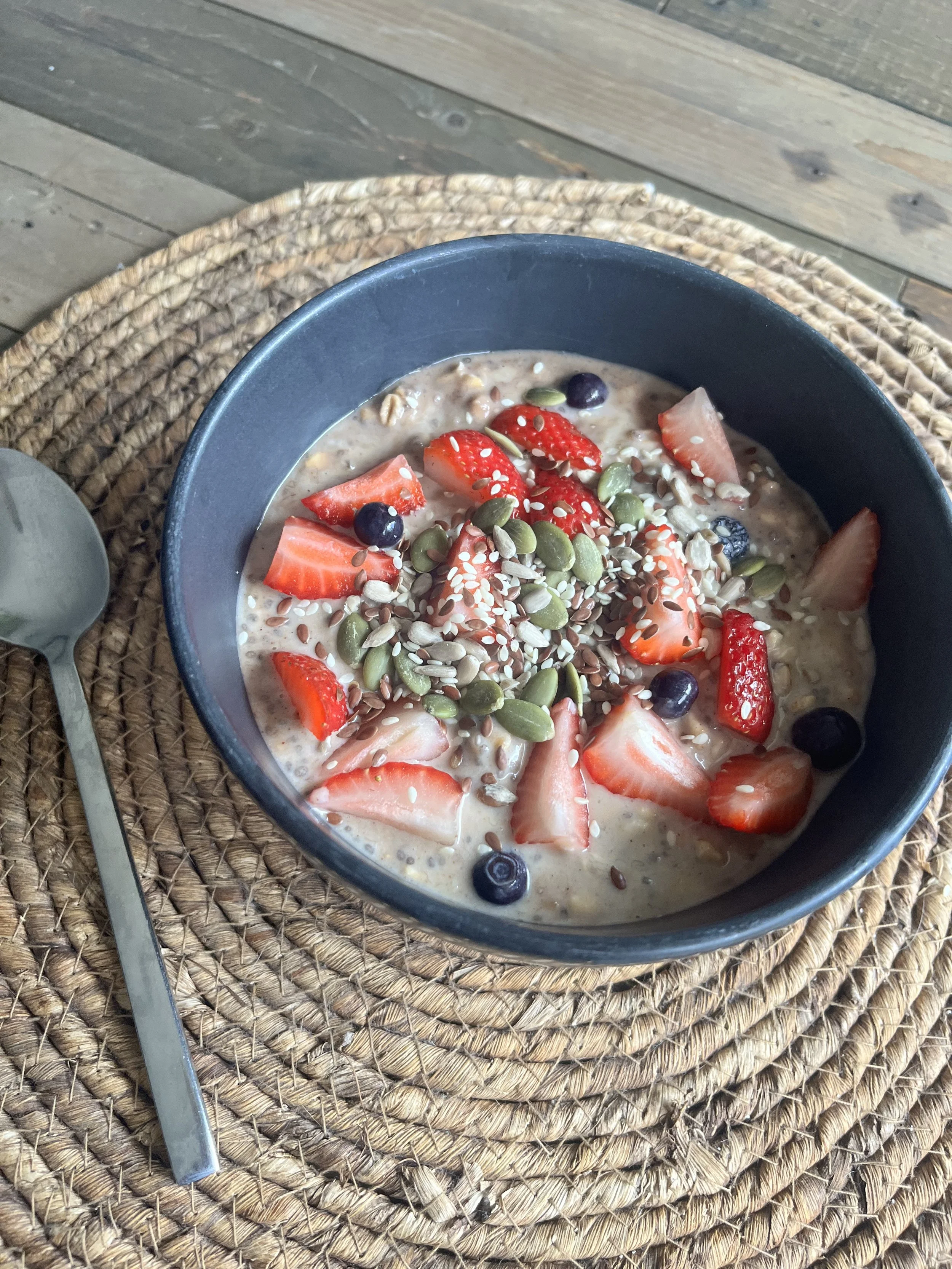 A black bowl filled with oatmeal topped with sliced strawberries, blueberries, pumpkin seeds, sunflower seeds, and sesame seeds, placed on a woven placemat on a wooden table with a silver spoon beside it.