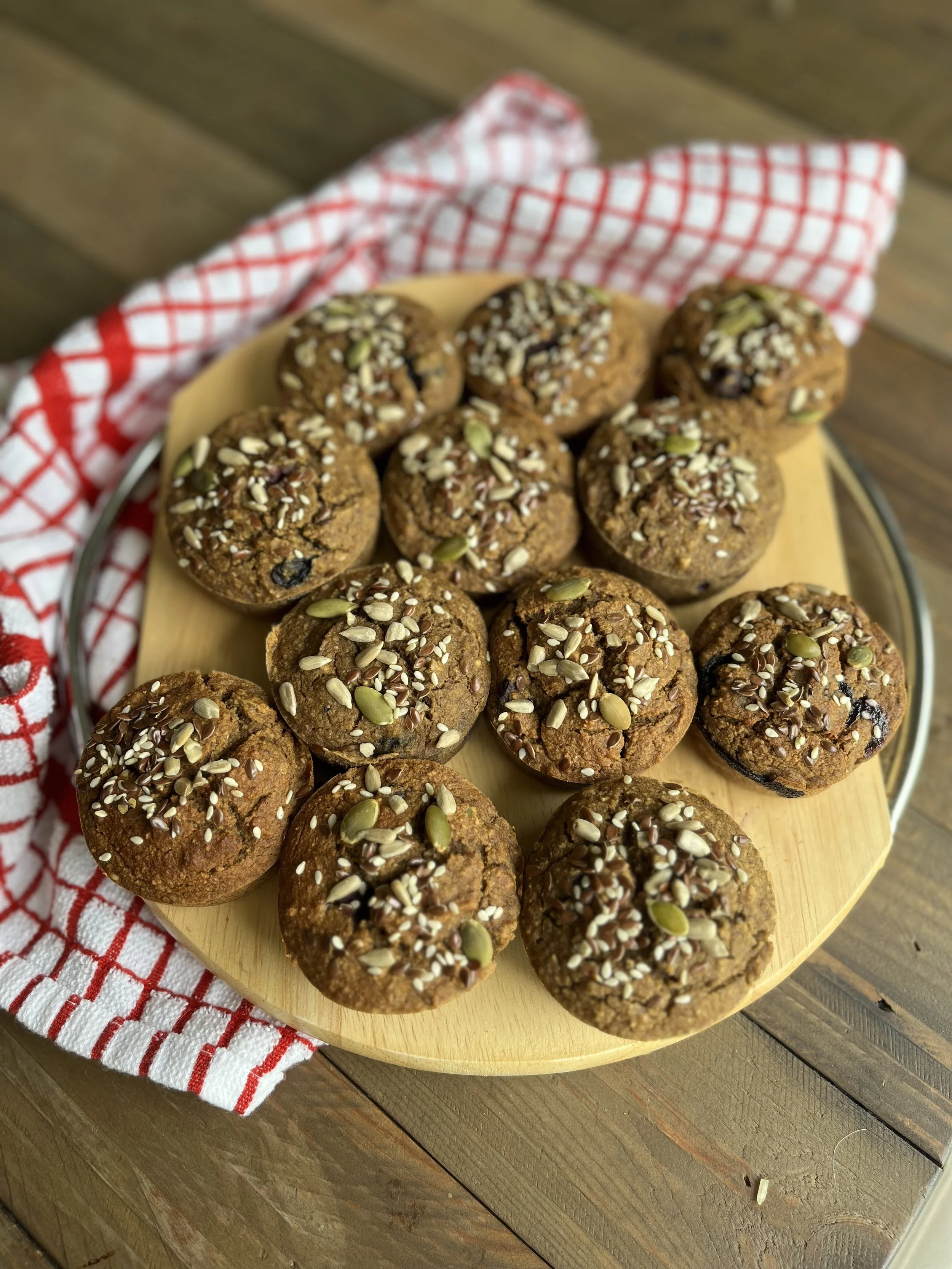 A plate of baked muffins topped with seeds, arranged on a wooden round tray with a red and white checkered cloth underneath.