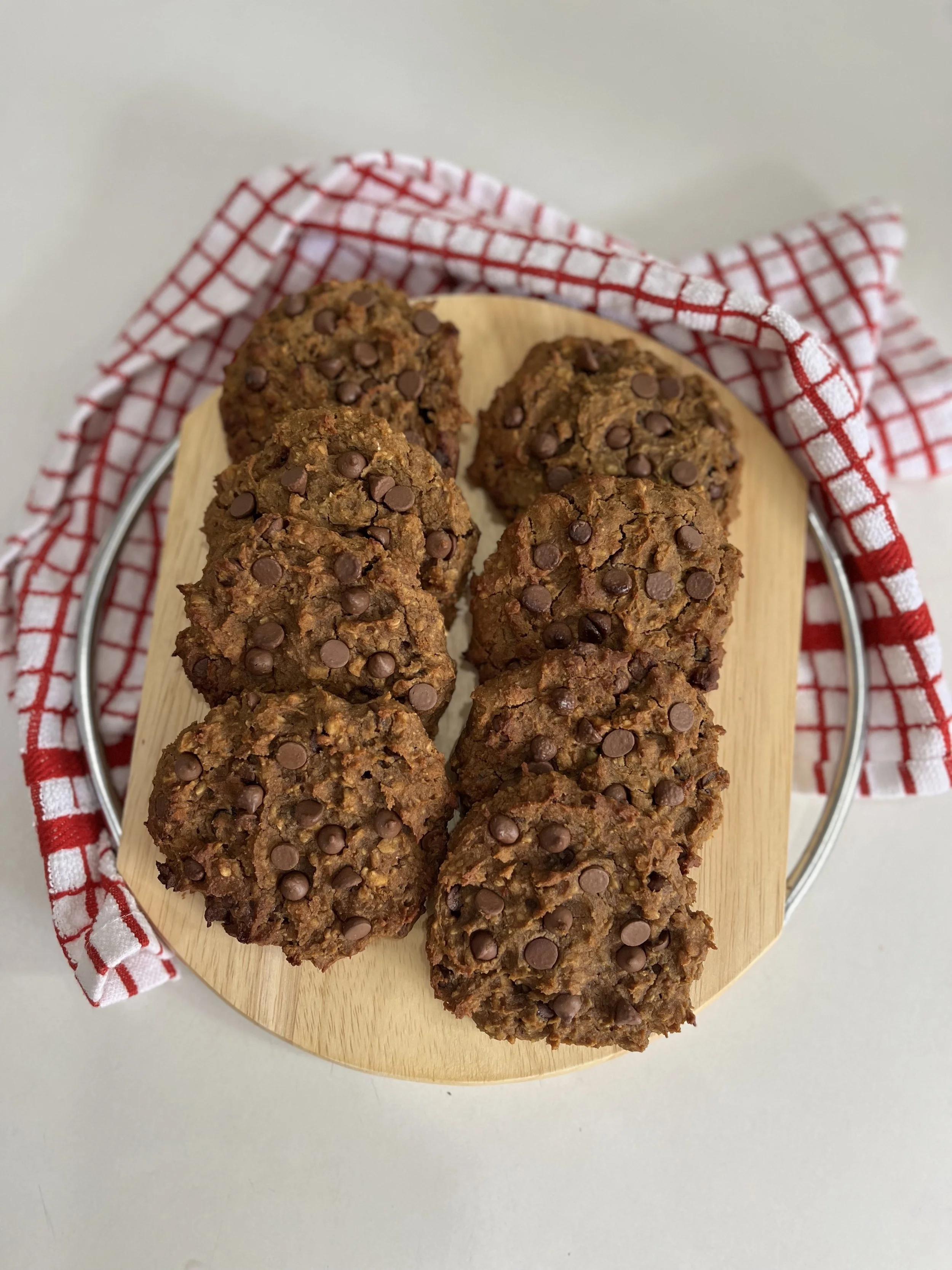 Six chocolate chip cookies on a round wooden tray lined with a red and white checkered cloth.