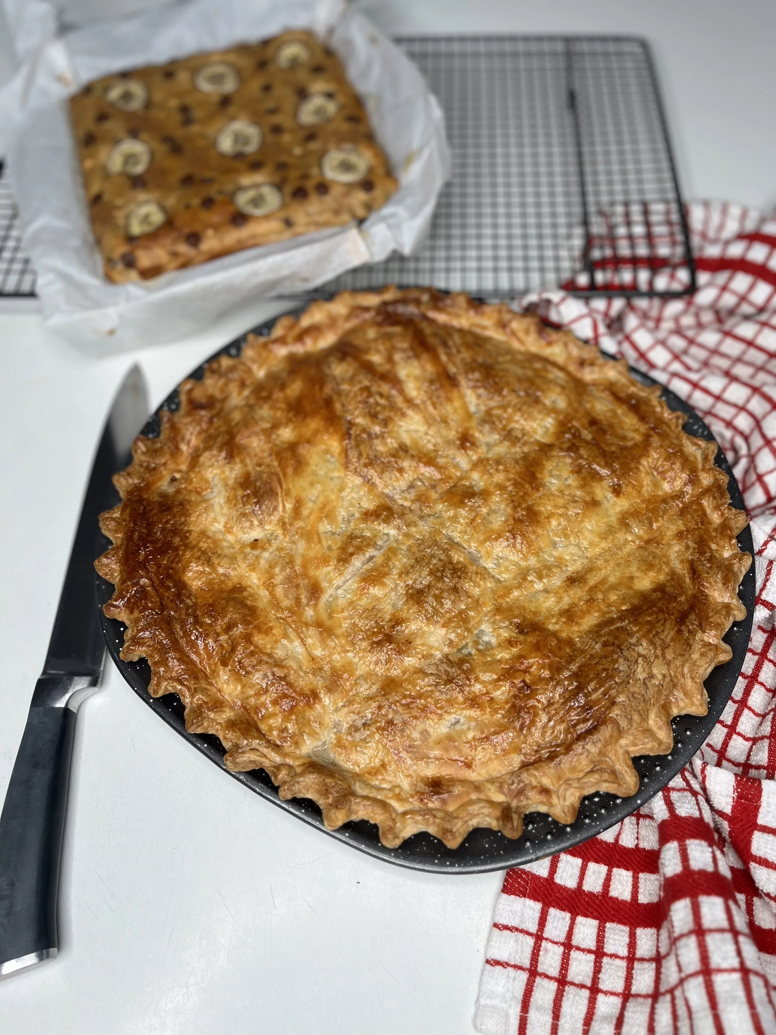 Hot homemade chicken pot pie on a black skillet with a golden brown crust, next to a kitchen towel with red and white checkered pattern, and a partially visible knife.