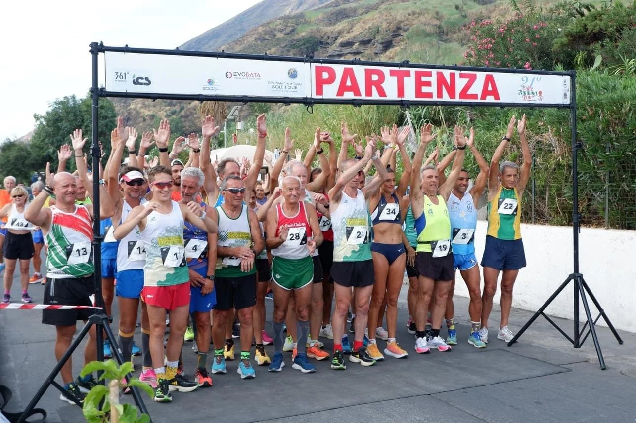 A group of runners at the starting line of a race, standing under a banner that says 'PARTENZA', with mountains and greenery in the background.