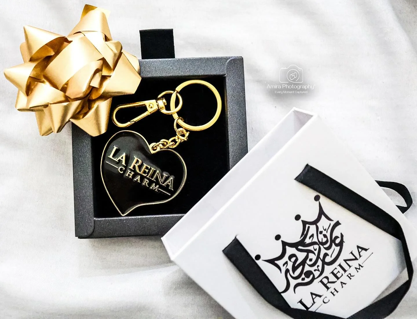 A black gift box with a gold bow, containing a LA REINA CHARM keychain, alongside a white box with black Arabic calligraphy and LA REINA CHARM branding.