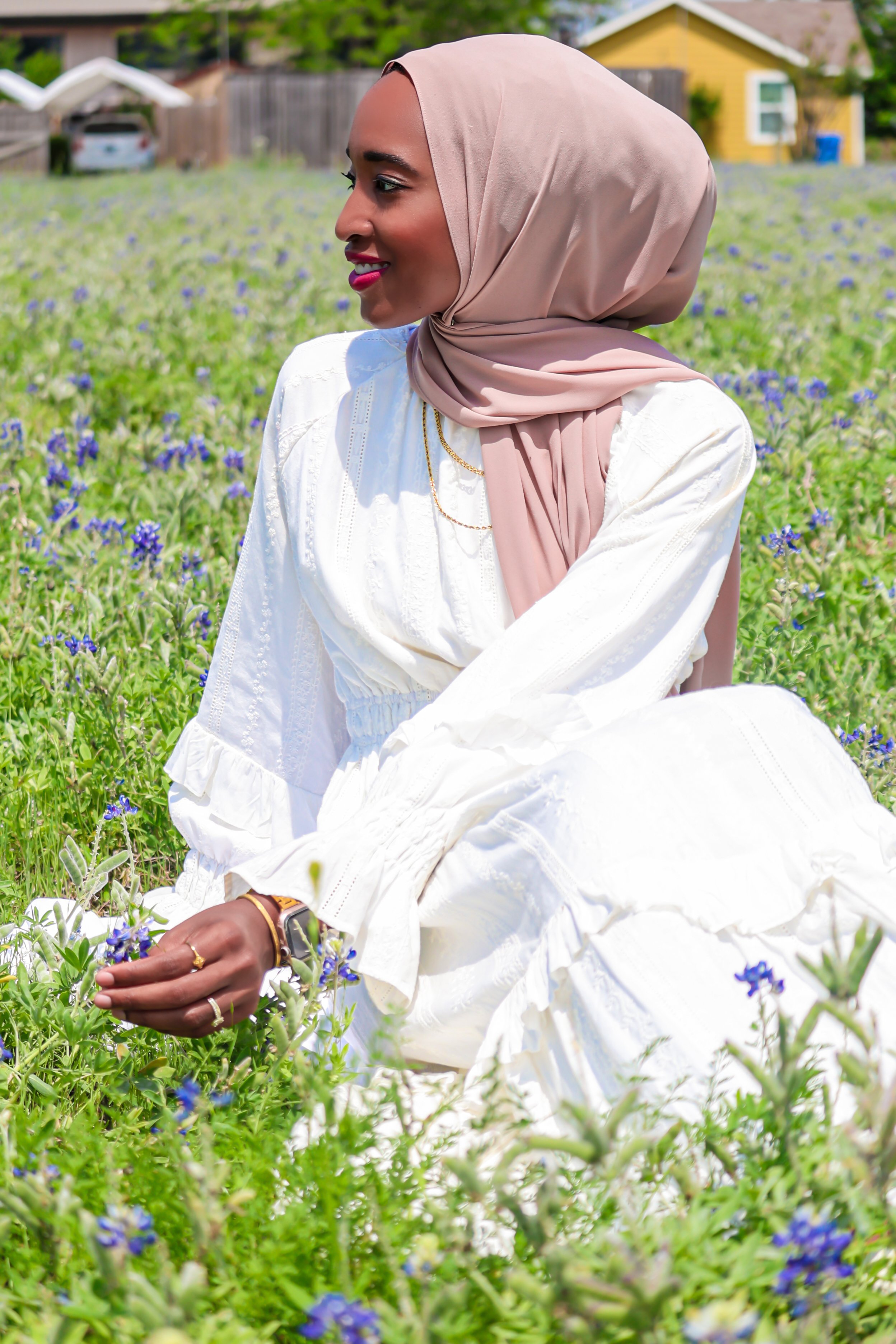 A woman in a beige headscarf and white dress sitting in a field of small blue flowers, looking to the side and smiling.