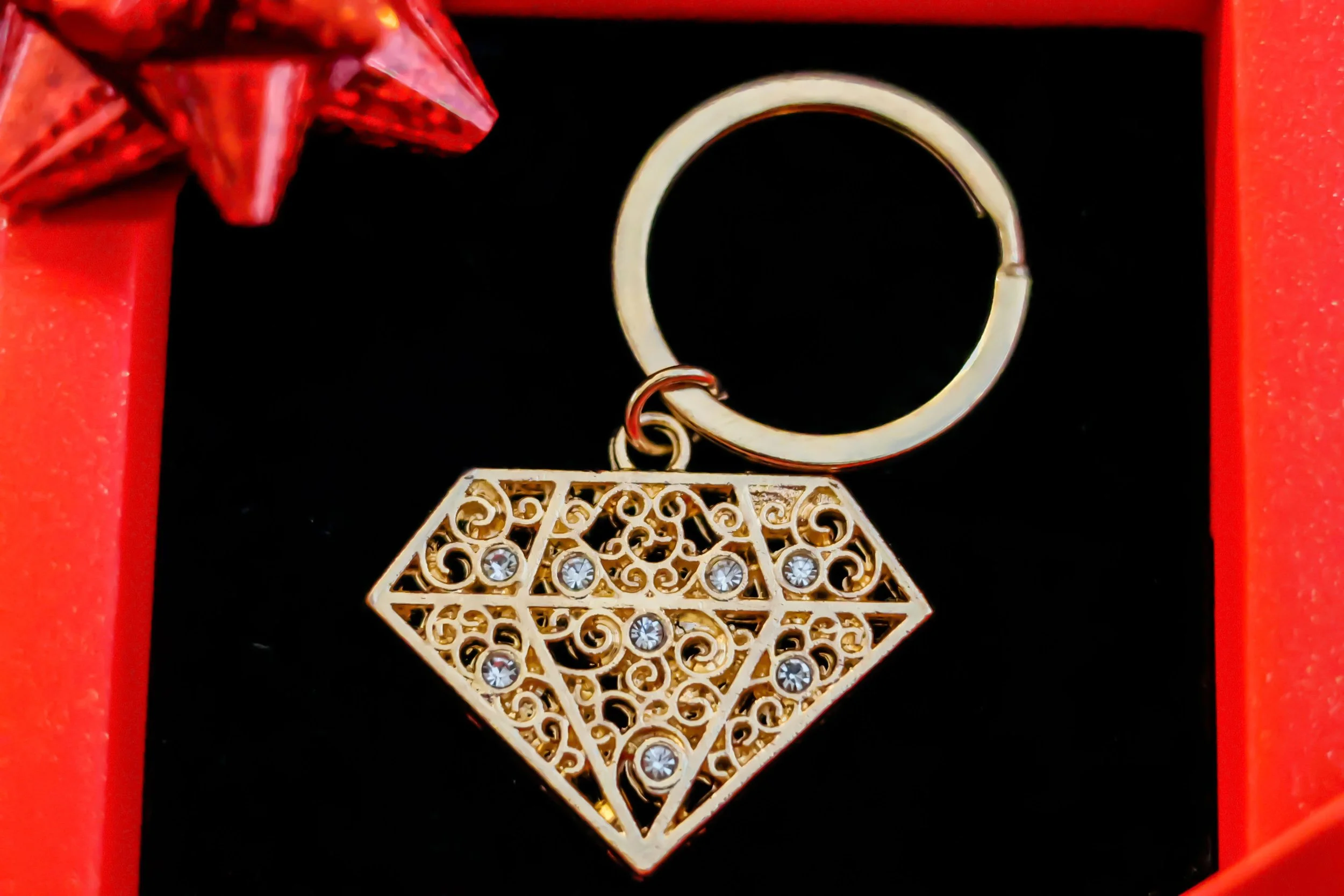 A gold-colored diamond-shaped pendant necklace with intricate filigree design and embedded clear gemstones, hanging from a keyring hook inside a red jewelry box.