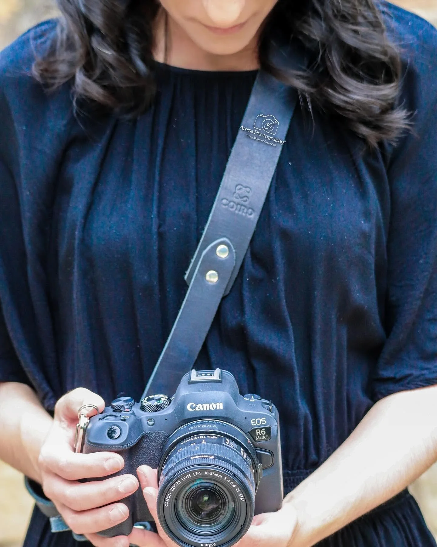 A person holding a Canon EOS camera with a zoom lens, wearing a dark blue top and a camera strap around their neck in a bright indoor setting.