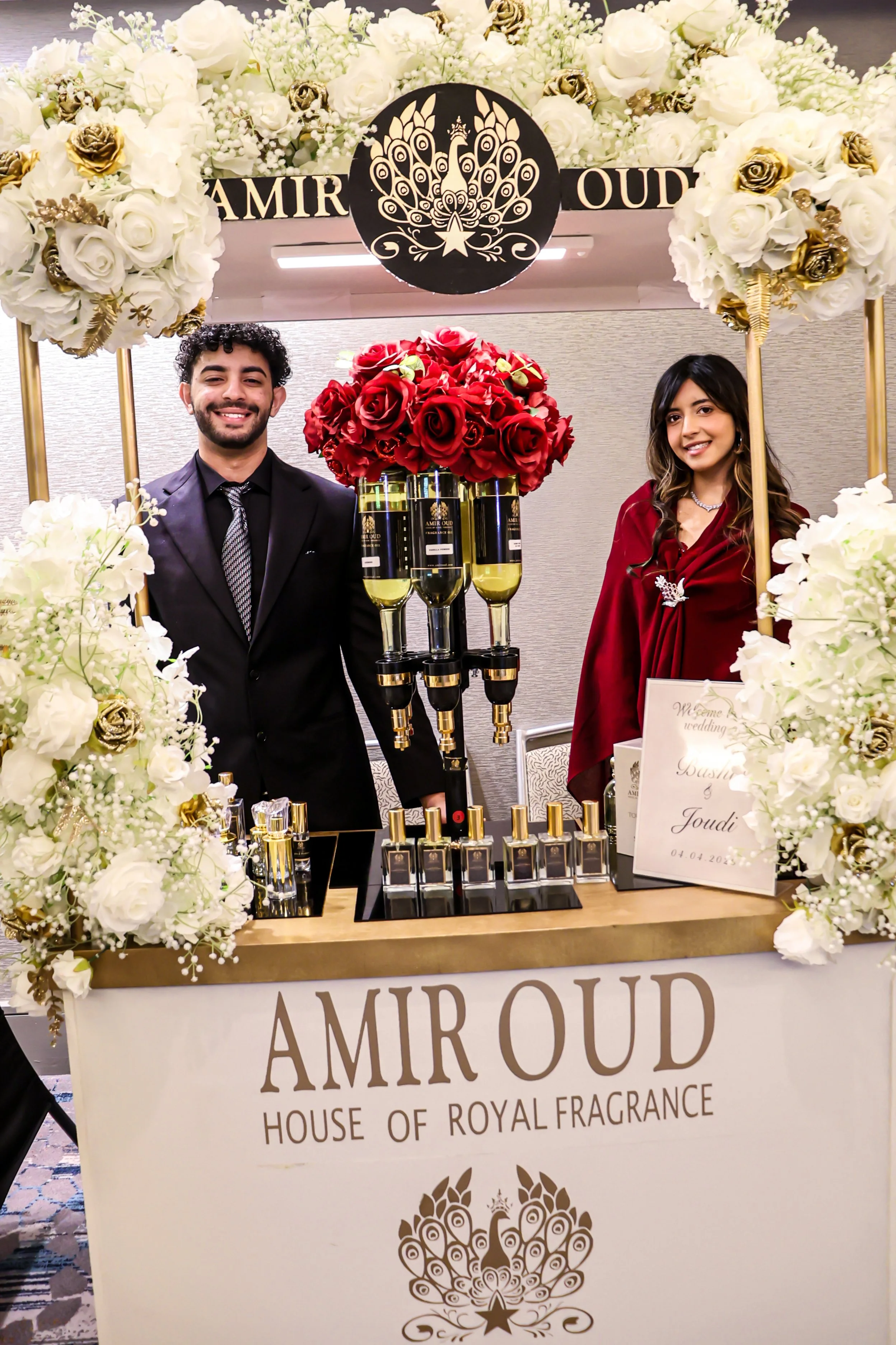 A floral arrangement featuring white and gold roses surrounds a display booth for 'Amir Oud,' a luxury house of royal fragrance. The booth has a central display with red roses, perfume bottles, and a sign with the event date, April 4, 2023, at a trade show or similar event.