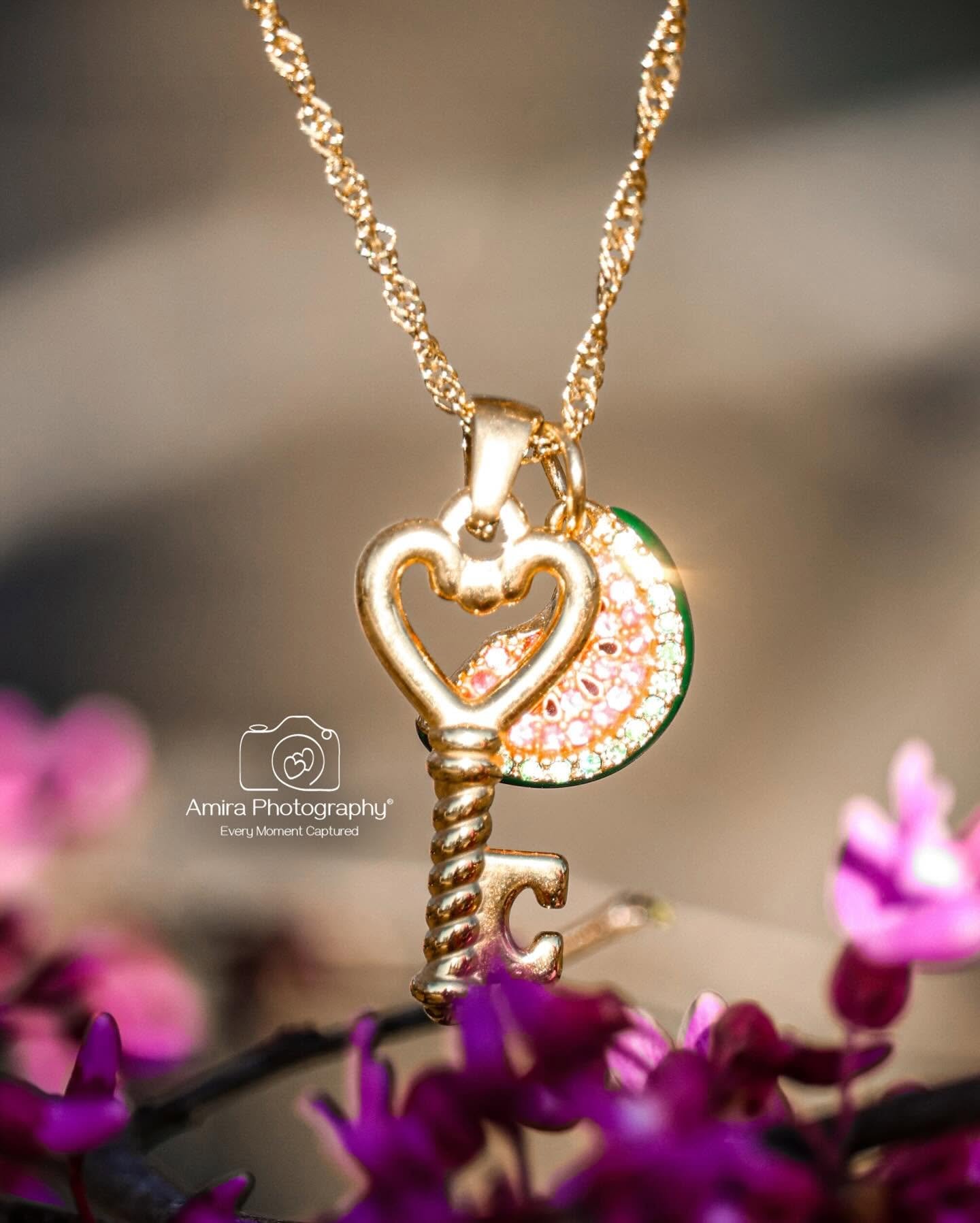 Close-up of a gold necklace hanging with a key and heart-shaped pendant, surrounded by purple flowers.