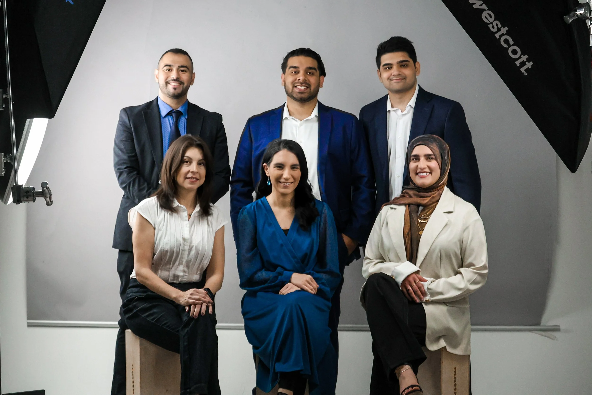 A diverse group of six professionally dressed people, three men standing in the back and three women sitting and standing in front, posing for a photo in a studio with lighting equipment visible on the sides.