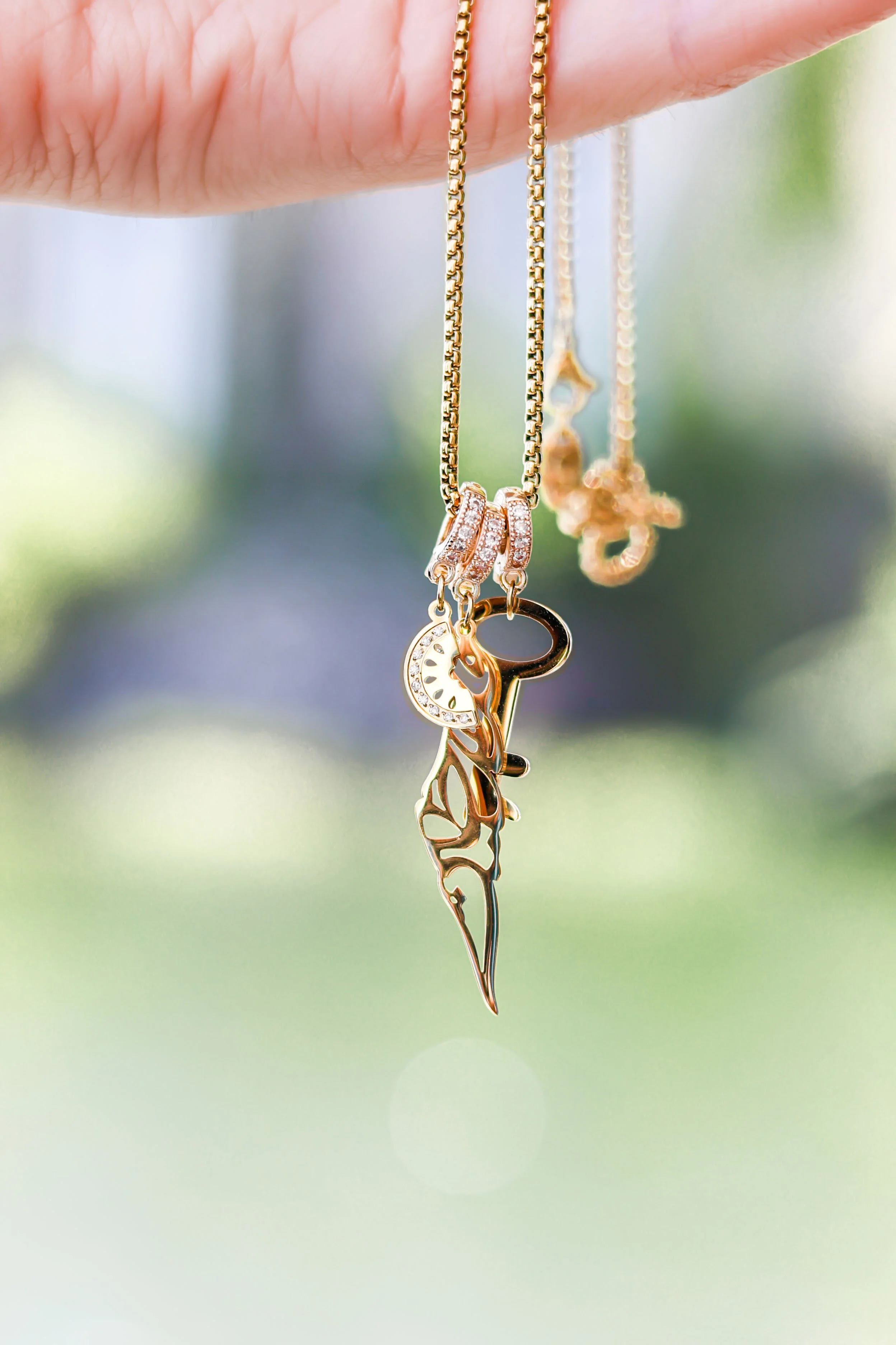 Close-up of a hand holding three gold necklaces with pendants, set against a blurred green background.