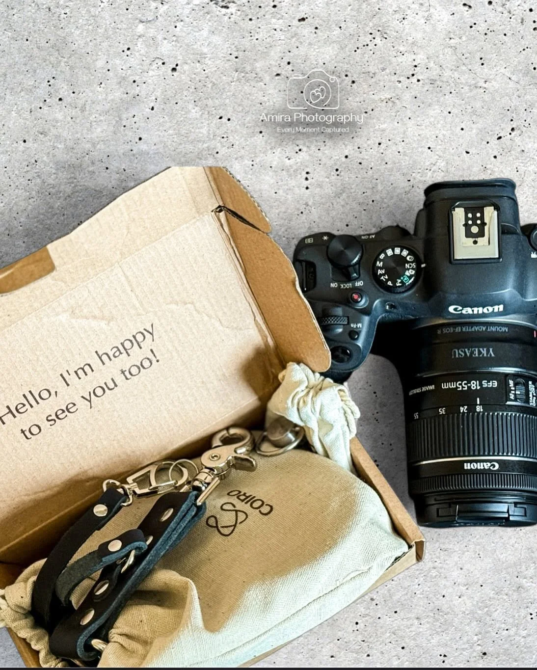 A camera and camera accessories in an open cardboard box on a speckled concrete surface, with a message inside the box reading 'Hello, I’m happy to see you too!'