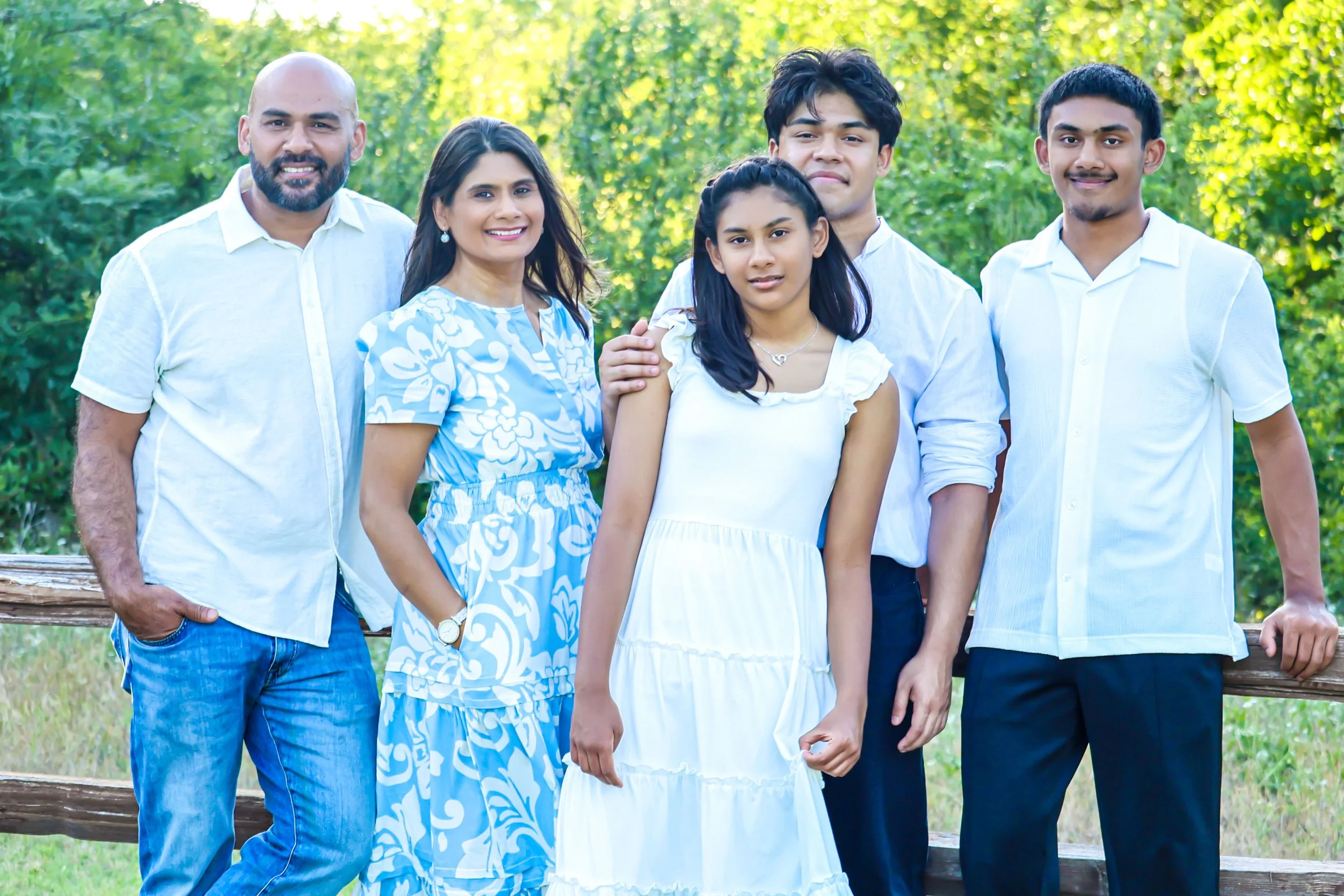 A diverse family of six people standing outdoors in a park, smiling for a group photo, with green trees in the background.