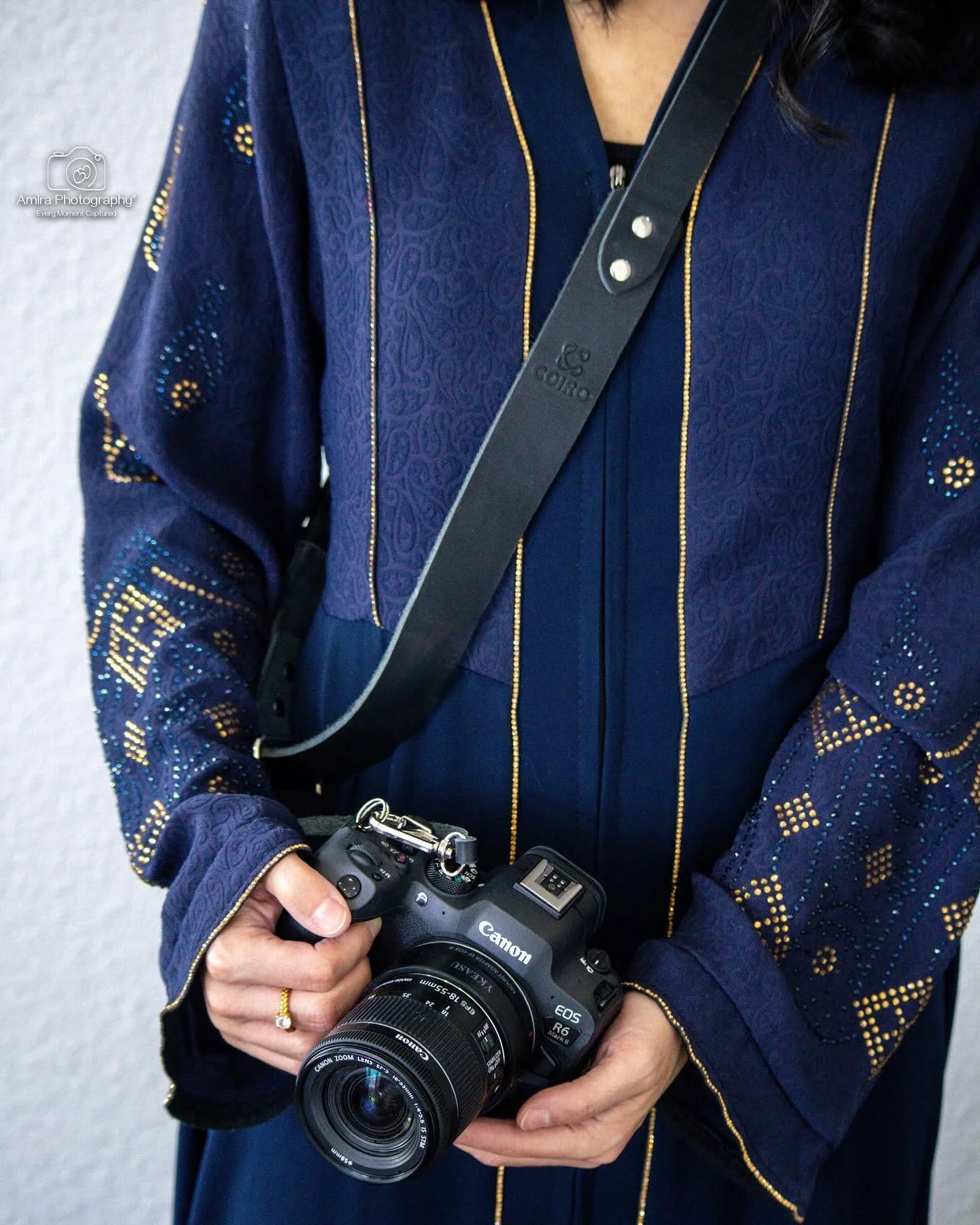A person wearing a blue embroidered jacket with gold accents holding a Canon digital camera with their right hand, with a black camera strap across their chest.