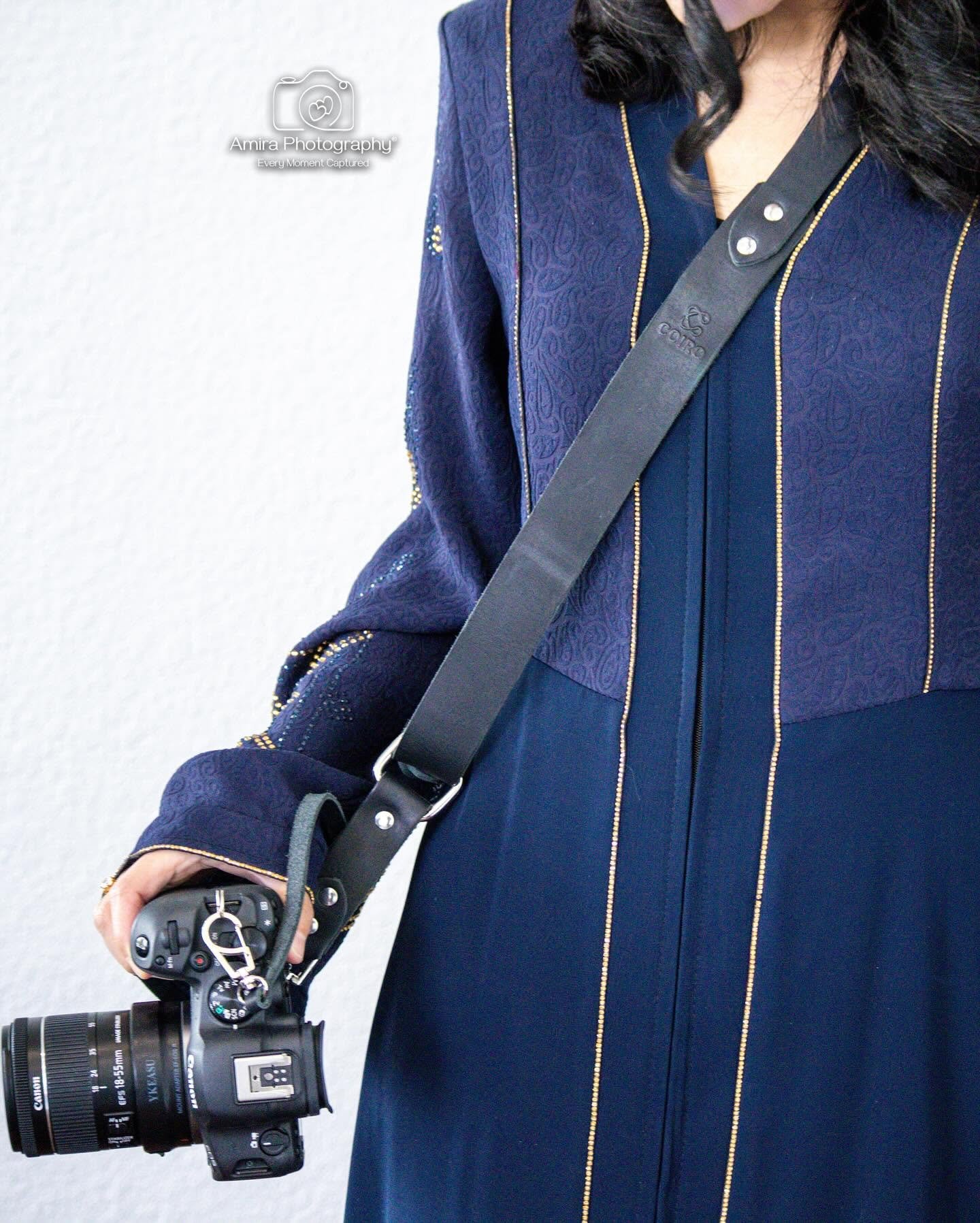 A woman with black hair wearing a navy blue jacket with gold trim, holding a camera with a long zoom lens, and wearing a camera strap that says 'COIRO' on it.