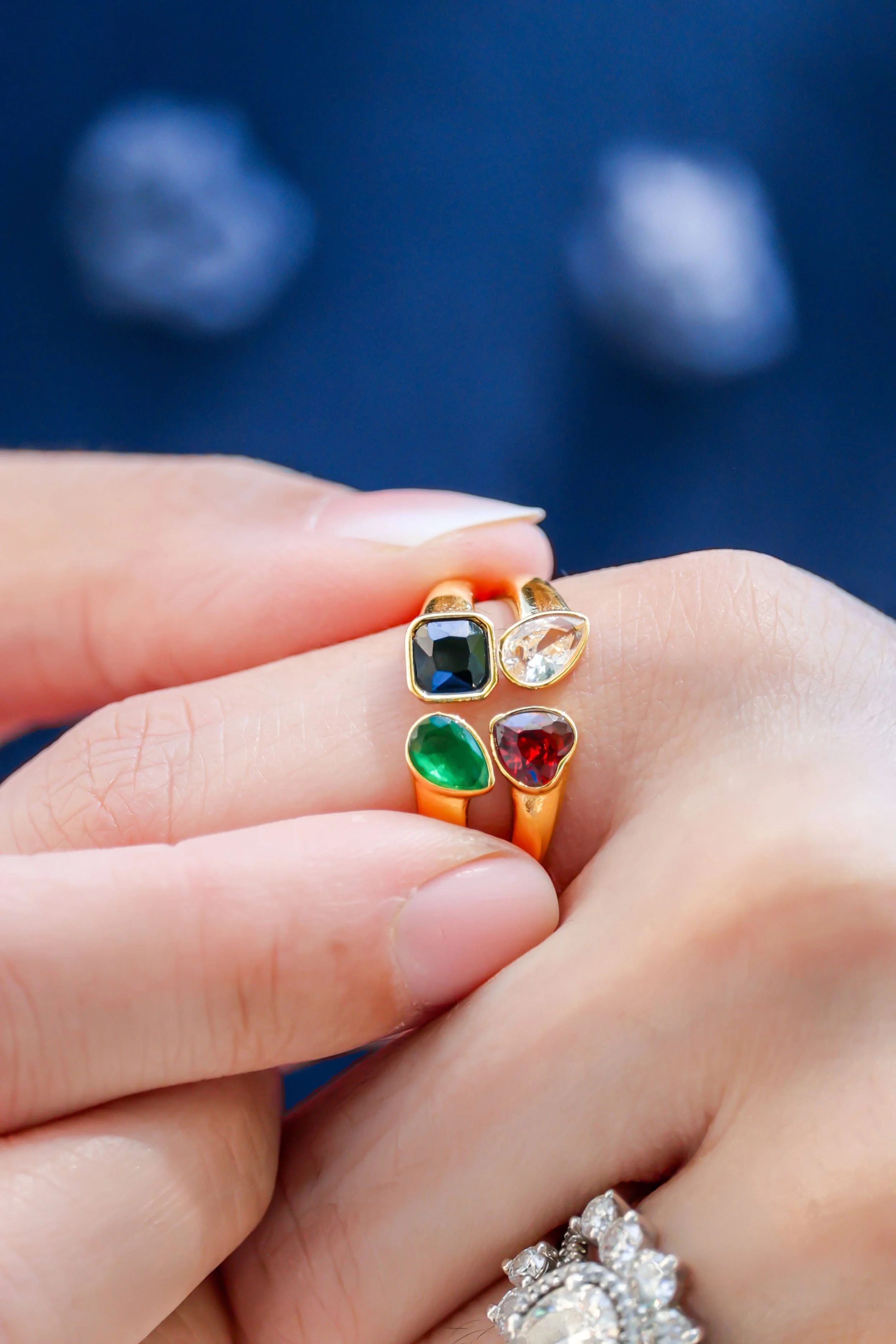 A person's hand wearing multiple rings, including a gold ring with a dark gemstone, a clear gemstone, a green gemstone, and a red gemstone. The person is holding their fingers close together, and other rings are visible on their fingers. The background is dark blue.