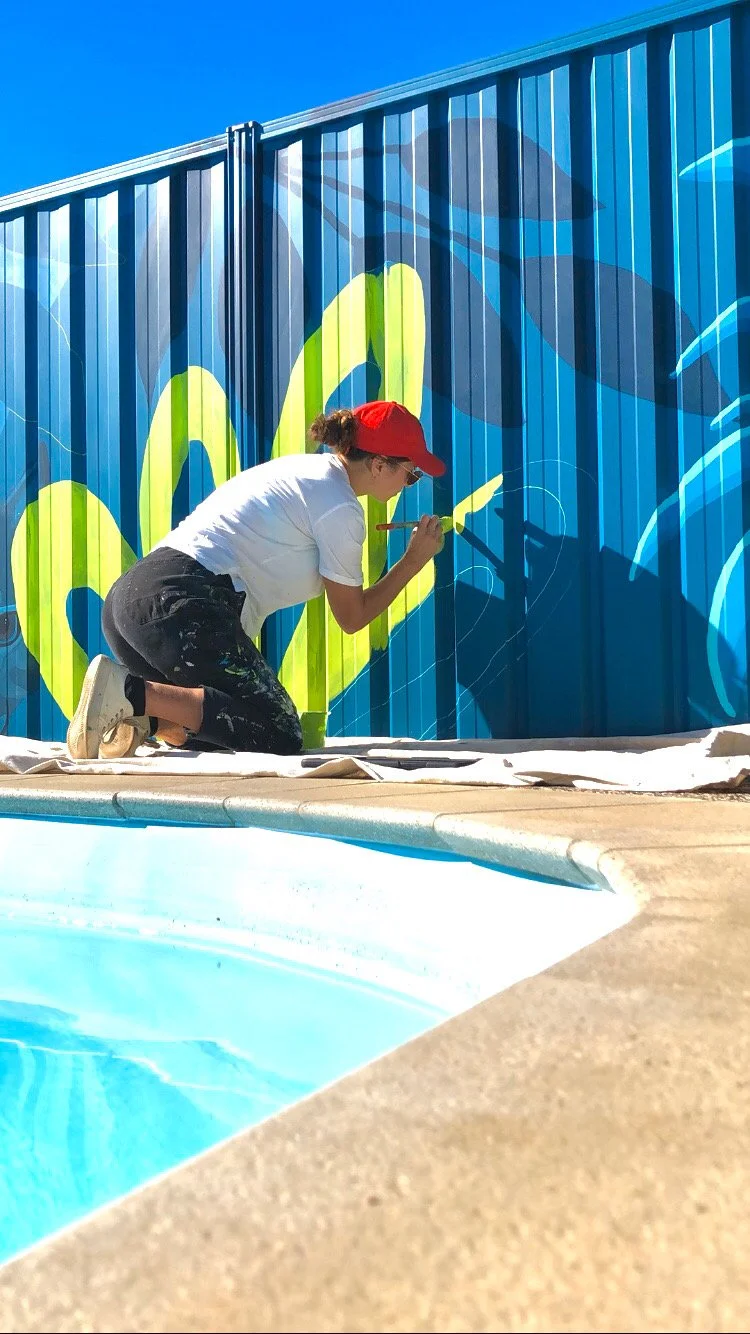 Person wearing a red cap and white shirt painting a colorful mural on a blue wall next to a swimming pool.