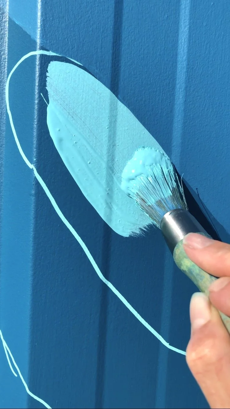 Close-up of a hand painting a light blue brushstroke on a dark blue surface with a paintbrush.