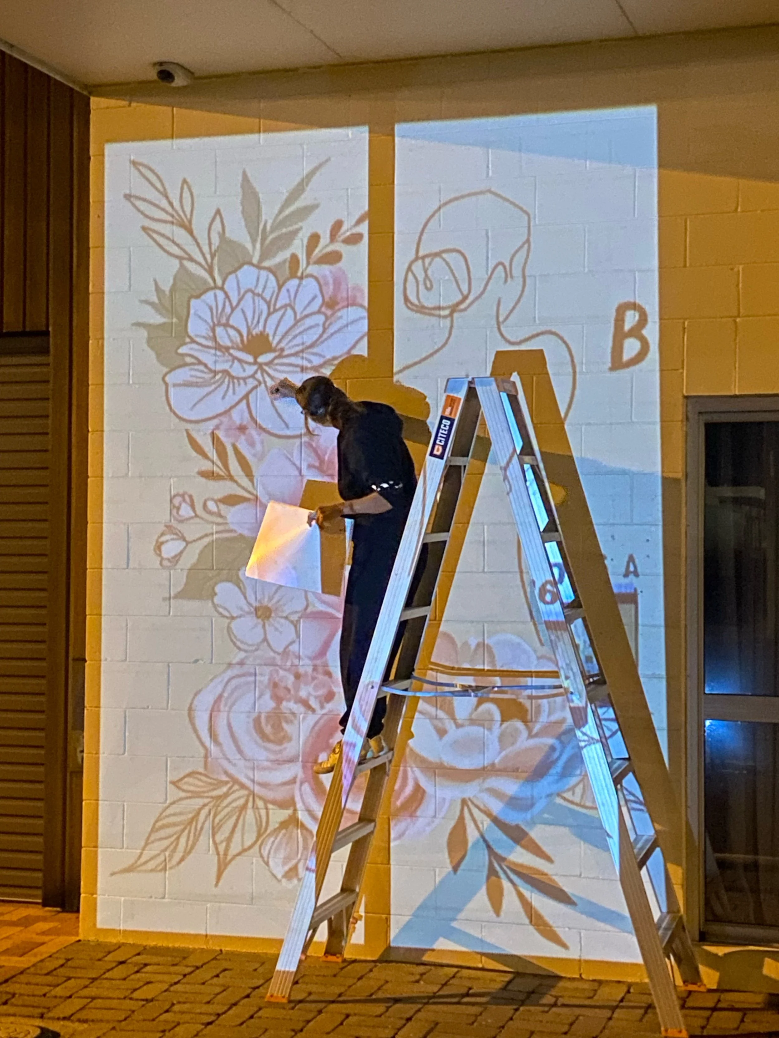 Person on ladder painting a mural with floral and abstract designs projected on a wall.