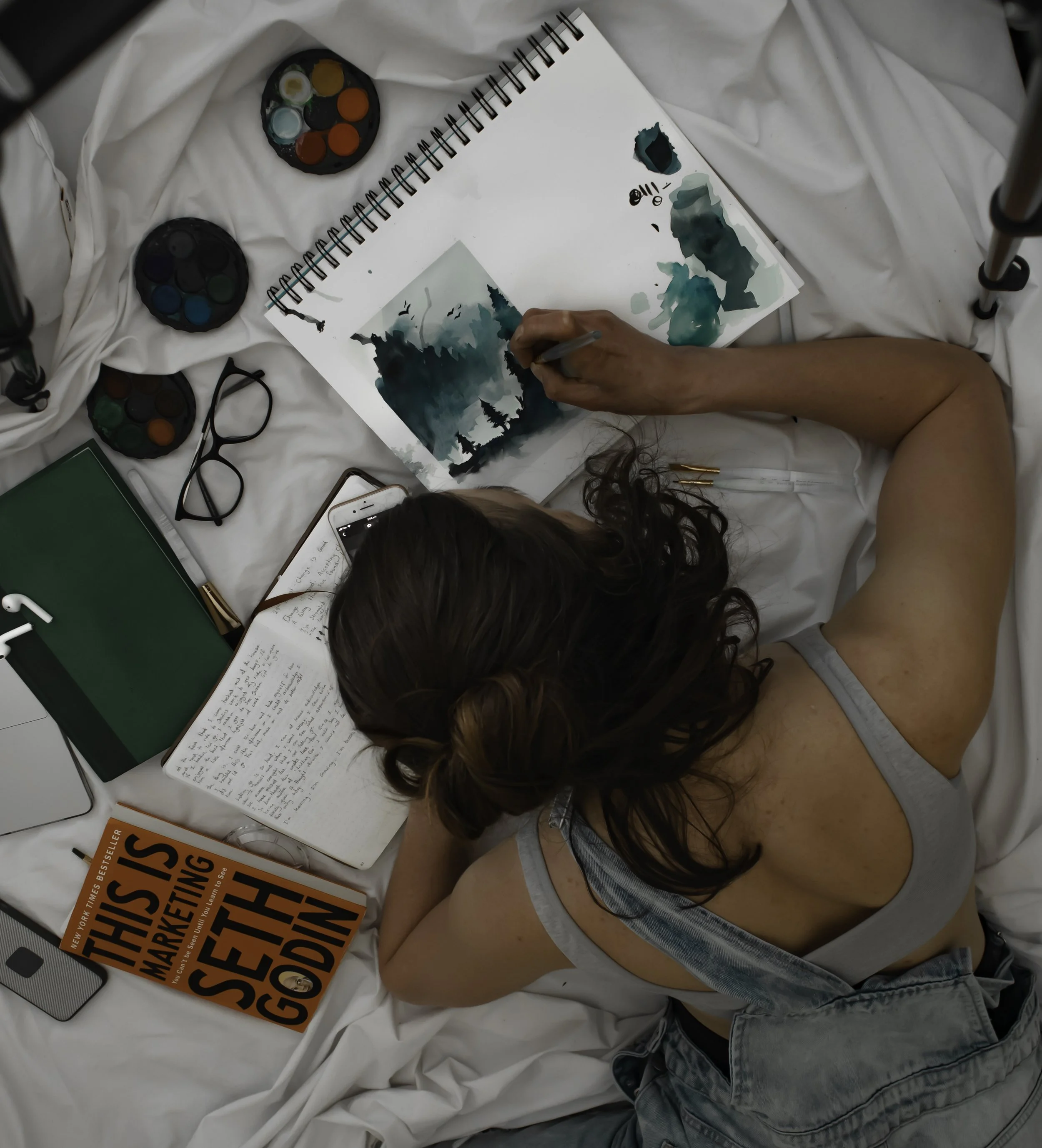 A person lying on a bed painting with watercolors in a sketchbook, surrounded by art supplies, a book titled 'This is Marketing' by Seth Godin, a notebook, glasses, a smartphone, and a laptop.