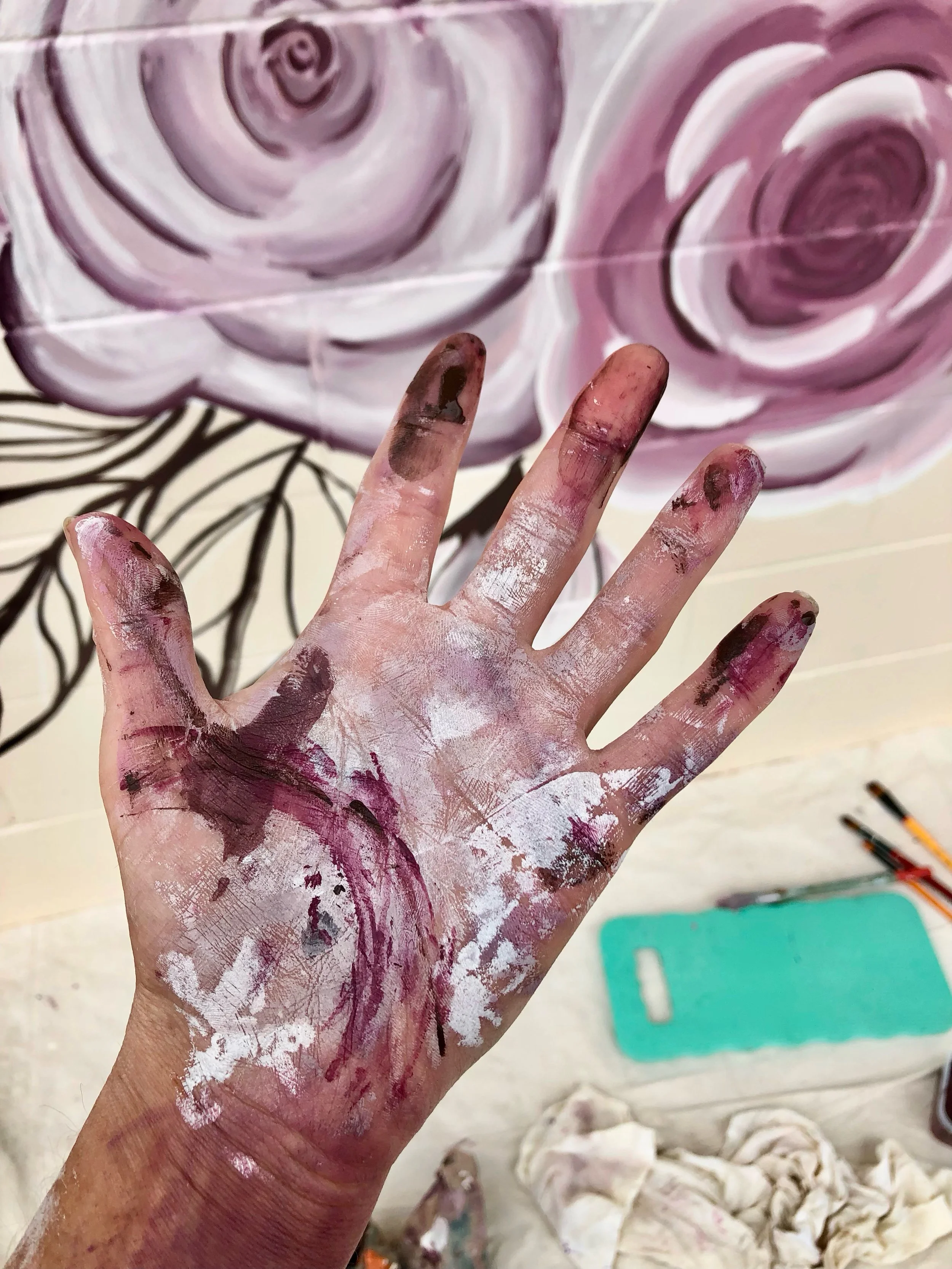 Artist's hand with paint smudges against a mural background of roses and leaves, paintbrushes, and painting supplies visible.