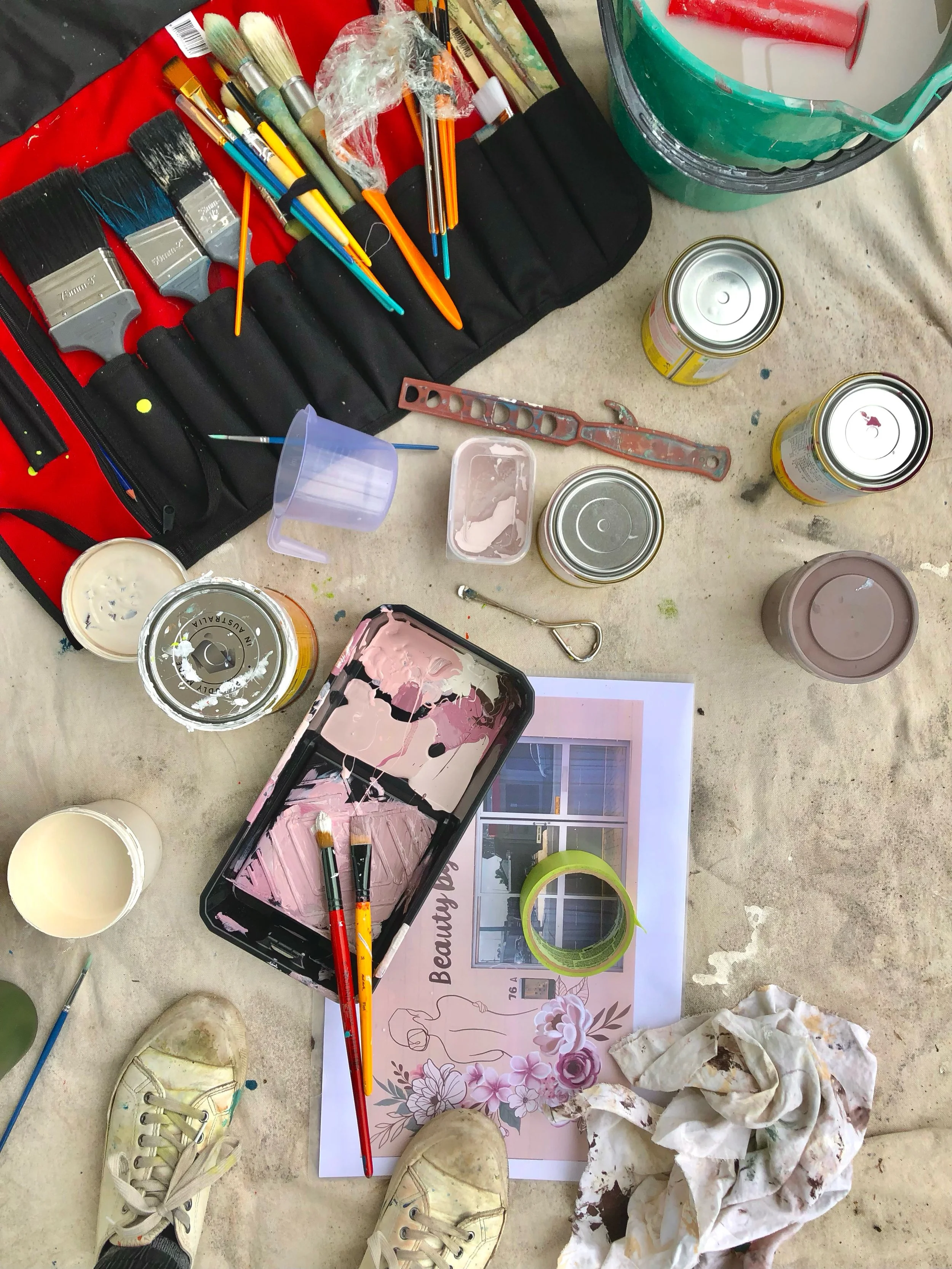 Assorted painting tools and supplies on a floor, including paintbrushes, paint cans, a paint tray with mixed colors, a paper with floral design, and a pair of shoes.