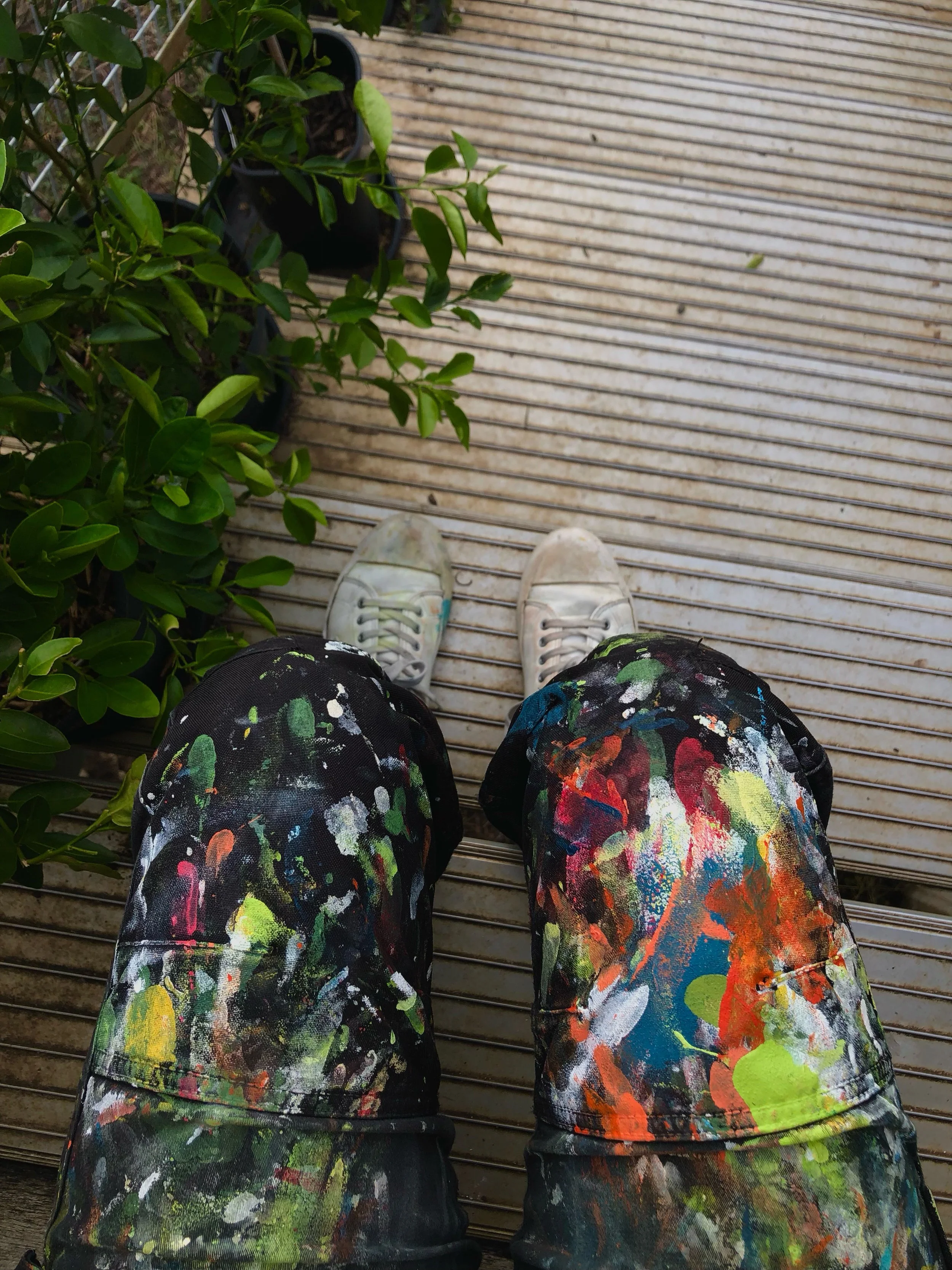 Person wearing paint-splattered pants, white sneakers, standing on wooden decking next to a leafy plant.