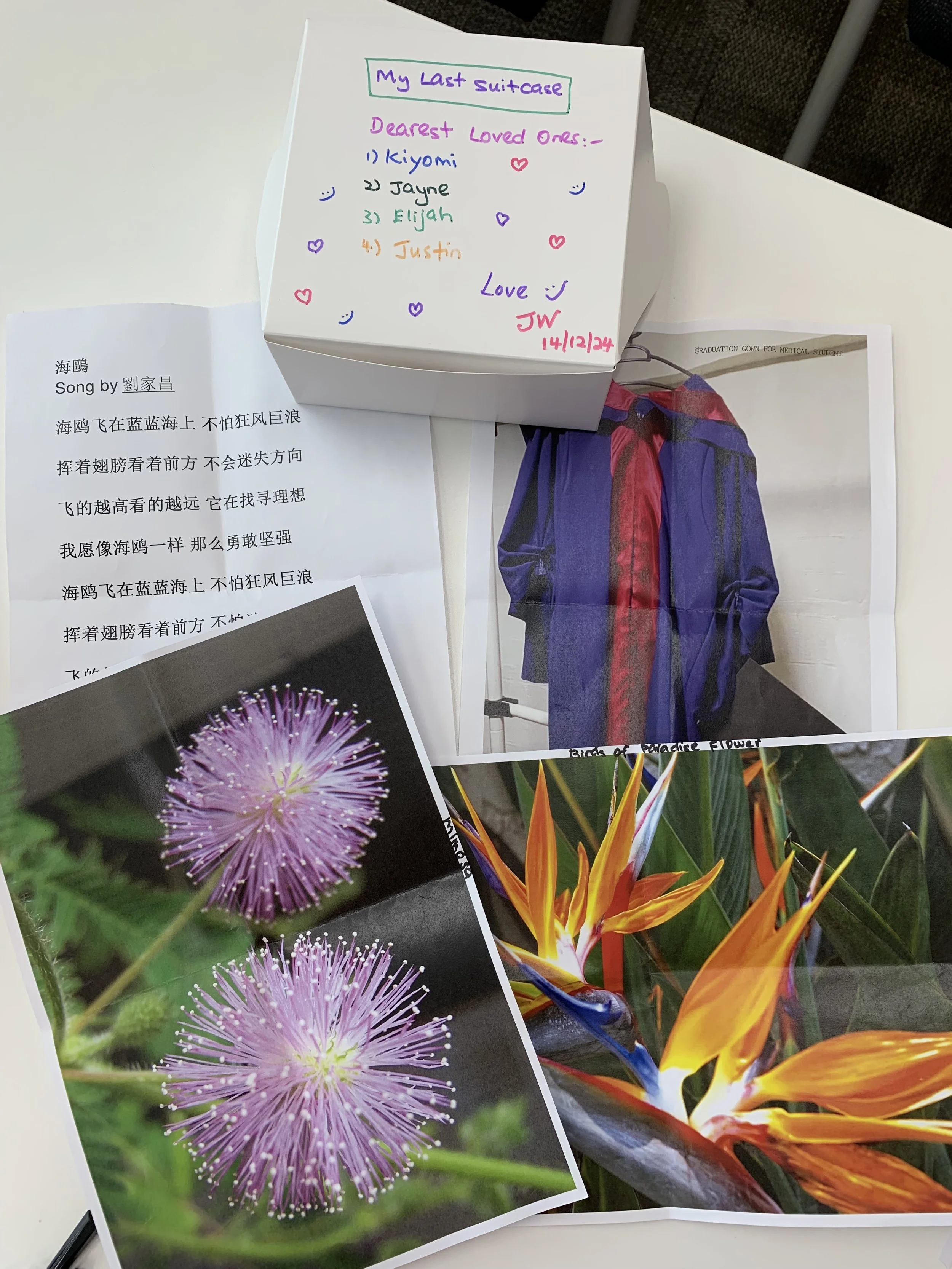A collection of papers and photographs on a table, including a handwritten note on a white box with colorful text and hearts, a page with a Chinese song, a photograph of purple flowers, and a photograph of orange and yellow flowers.