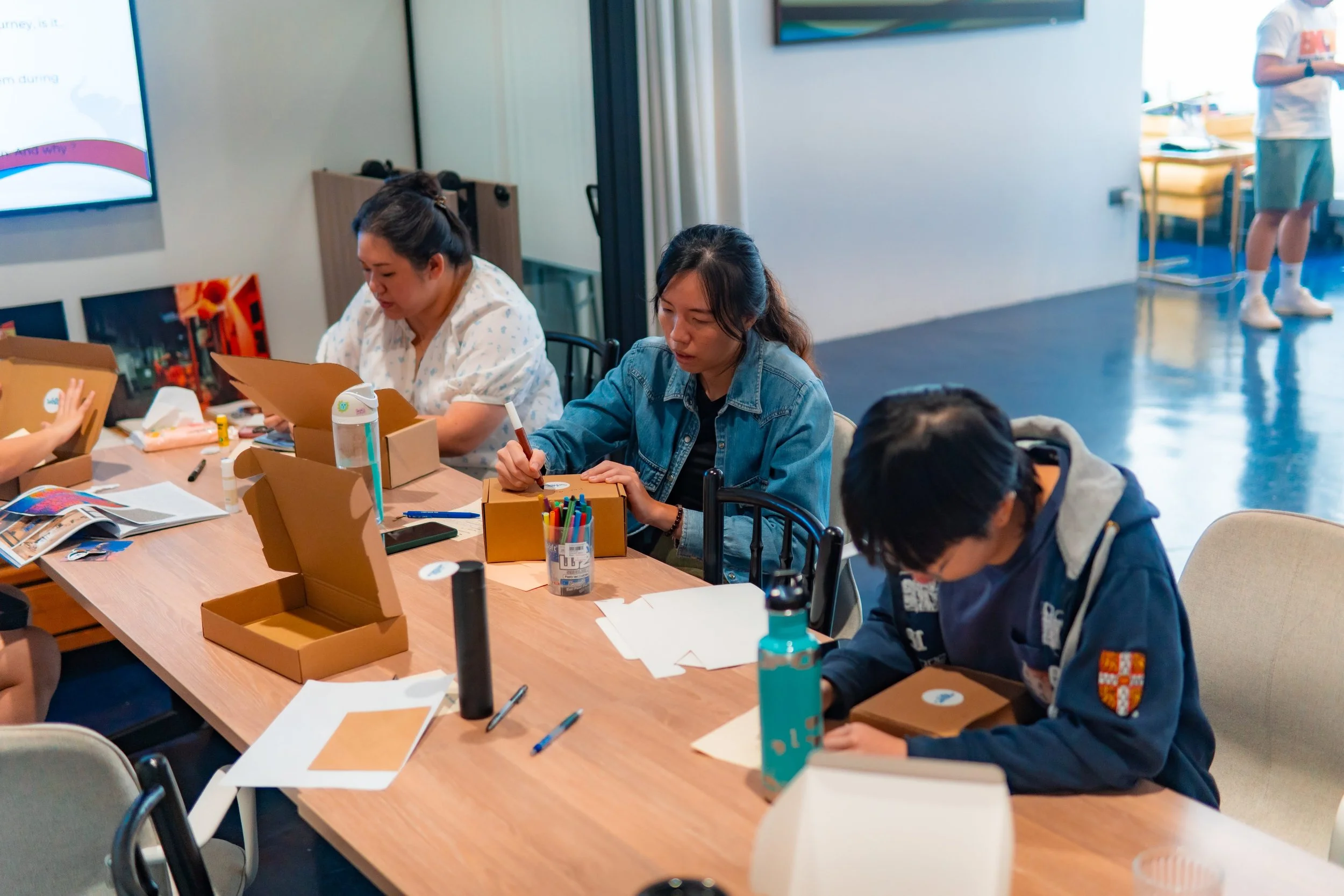 Three people sitting at a table engaged in craft activities with boxes, markers, papers, and art supplies