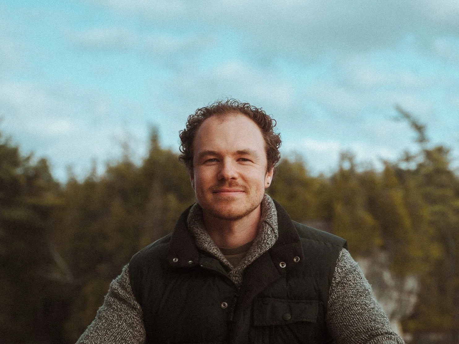 A man with curly hair and a slight smile, wearing a gray sweater and black vest, standing outdoors with trees in the background and a partly cloudy sky.