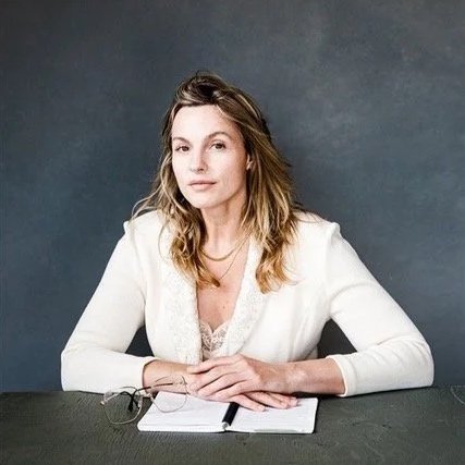 Woman with shoulder-length wavy hair wearing a white blazer, sitting at a table with a notebook and glasses, against a gray background.