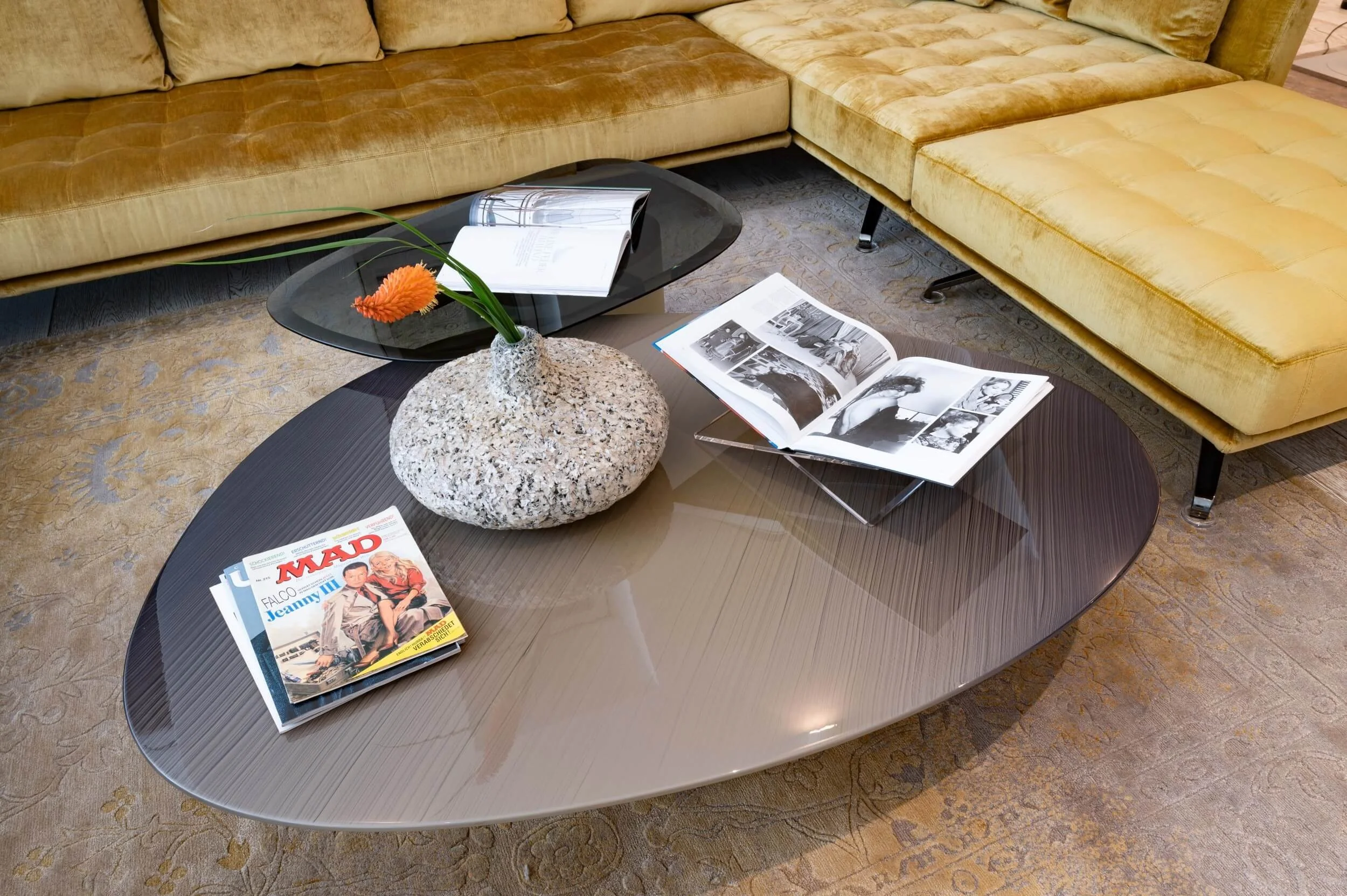 Detail der senfgelben Sofalandschaft mit organisch geformten Couchtischen, Teppich, Büchern und skulpturaler Vase im Wohnbereich.