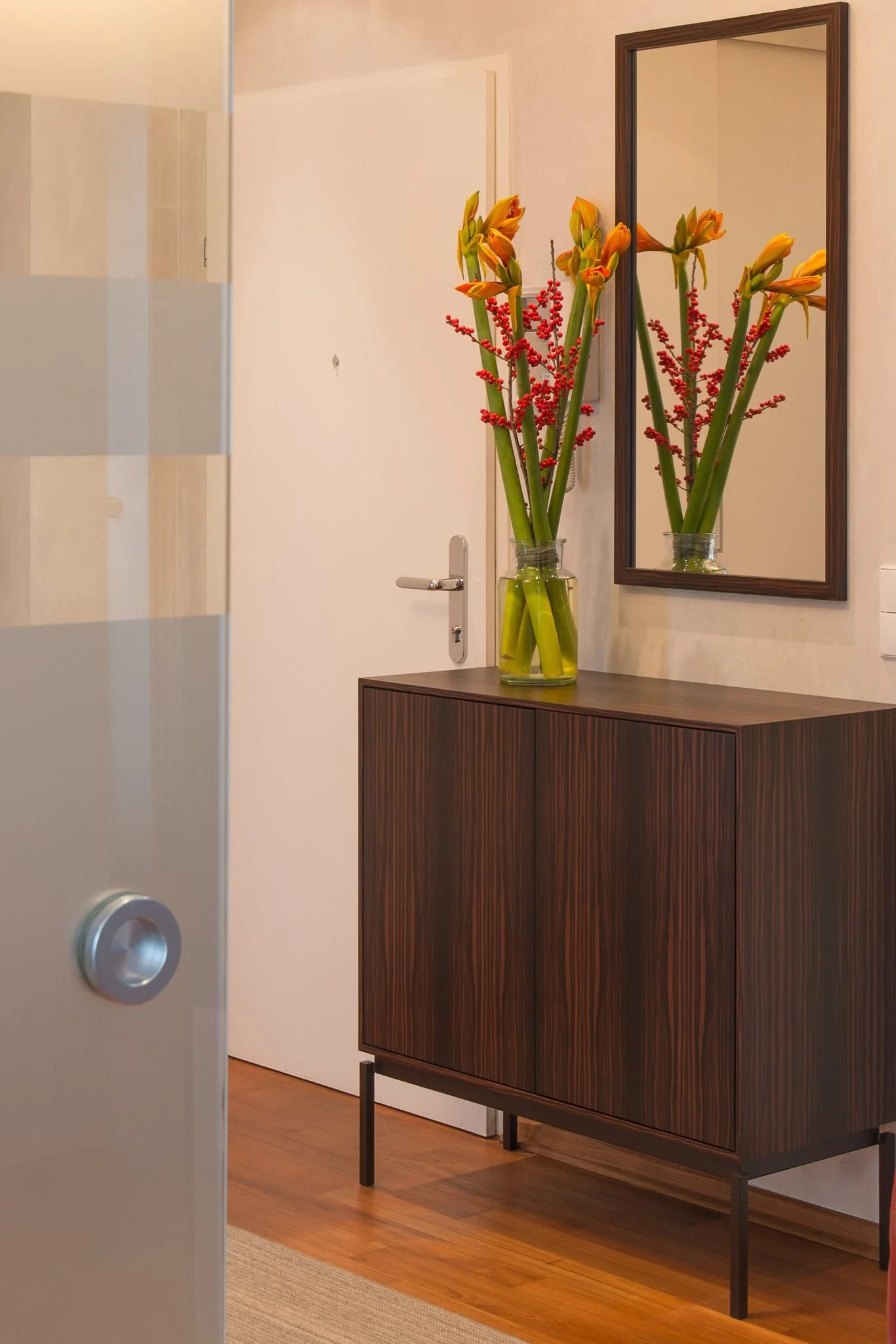 Elegantes Vorzimmer mit dunklem Holzschrank, gerahmtem Spiegel und dekorativem Blumenarrangement in Orange und Rot.