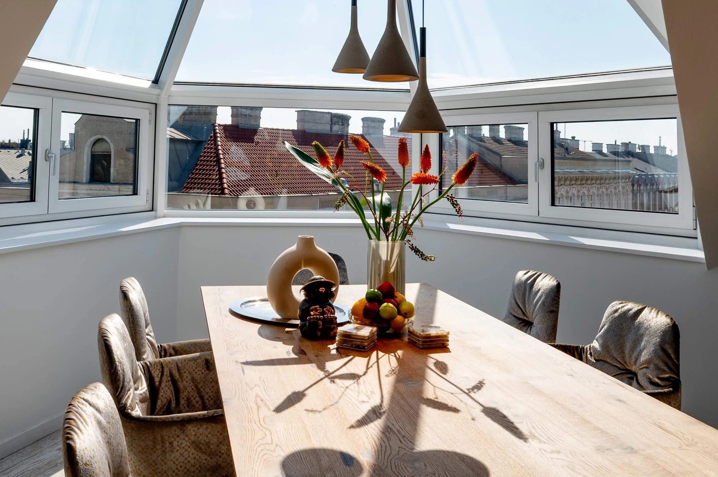 Detailaufnahme des Esstisches im sonnigen Dachgeschoss mit skulpturalen Pendelleuchten, floraler Dekoration und Blick auf die Wiener Dachlandschaft.